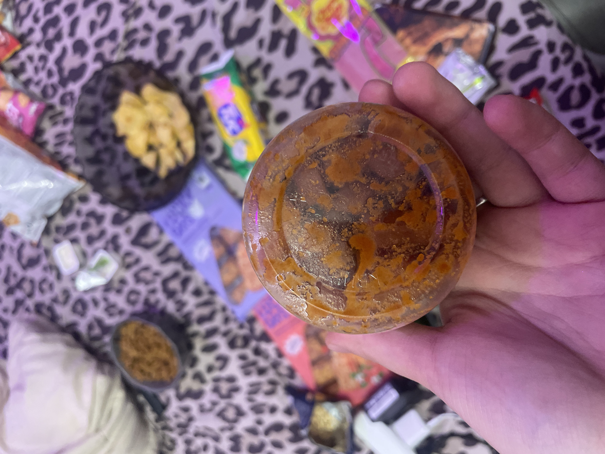A hand is holding a clear plastic container filled with a reddish-brown, chunky substance. The background is a leopard-print fabric, on which several snack food wrappers and containers are scattered. These include a dark bowl with yellow chips, a small container with what appears to be a white dip and green herbs, and various colorful packages. The scene is dimly lit, with pink and purple hues suggesting artificial lighting. The location is described as the city of Zmeinyi, Russia.