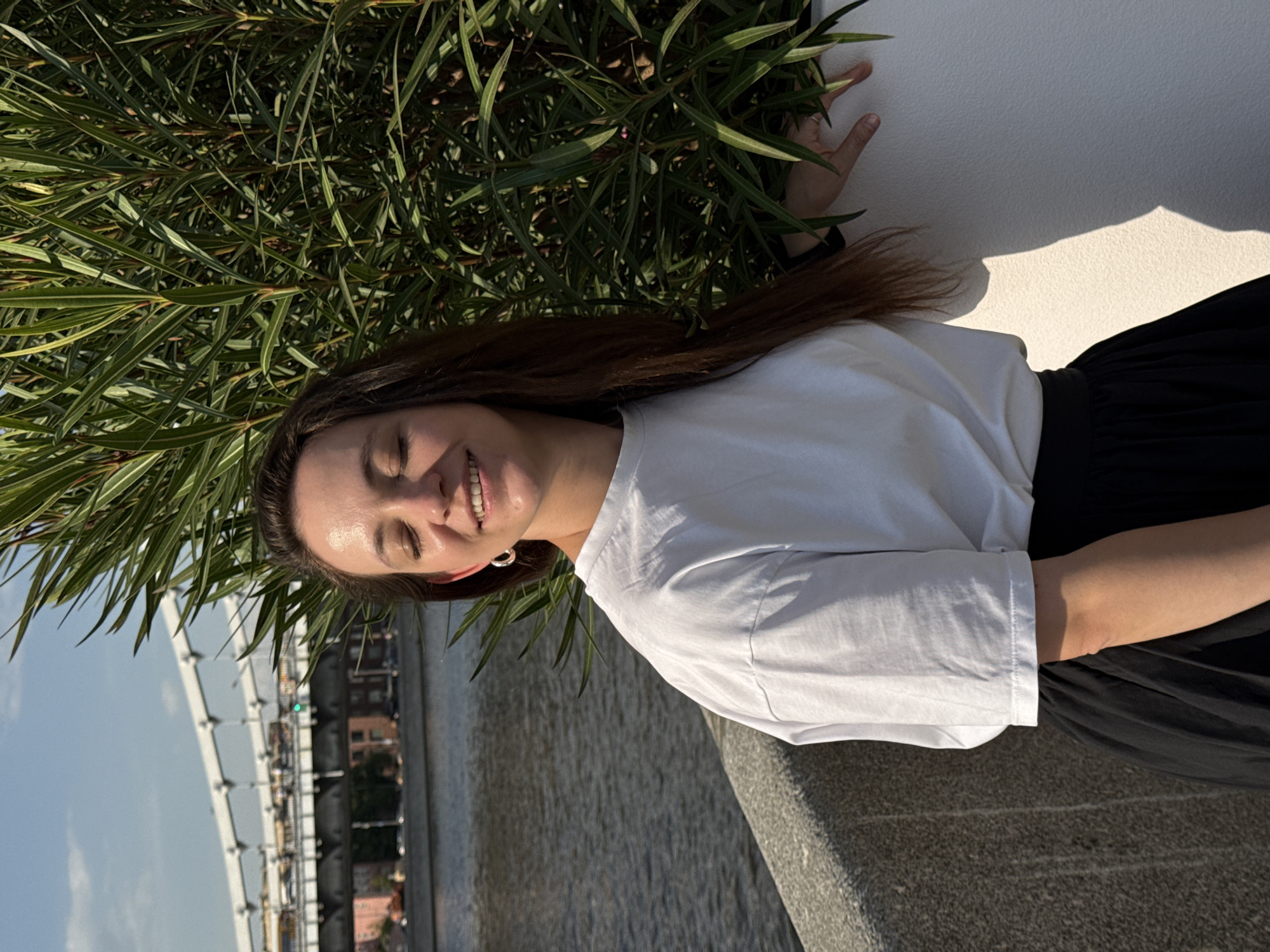 A woman stands next to a wall with green foliage in Moscow, Russia. Her brown hair is down, and she wears a white t-shirt and a black skirt. She is smiling with her eyes closed. A bridge, a river, and buildings are visible in the background. The wall she is standing next to appears to be made of concrete.