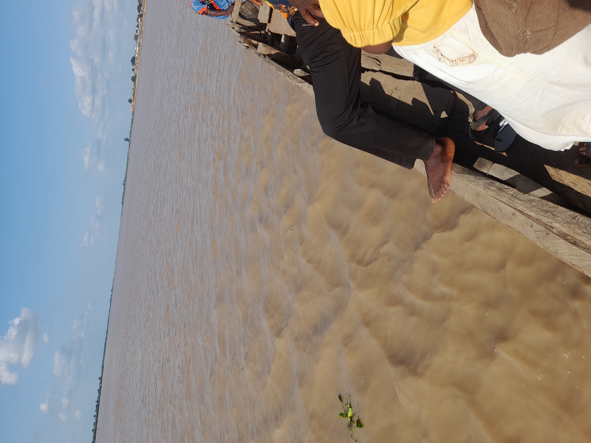 The image captures a view from a wooden boat on a wide, brown river, likely the River Benue near Ibi, Nigeria. The water is calm with gentle ripples. In the foreground, the legs and feet of several individuals are visible. One person is wearing black trousers and is barefoot. Another person is wearing white trousers and is partially visible. A third person, wearing a bright yellow shirt, is also in the frame. The boat itself is made of weathered wood. In the distance, the riverbanks are lined with sparse vegetation and what appear to be small structures, suggesting a settlement. The sky is blue with scattered white clouds, indicating a clear day.