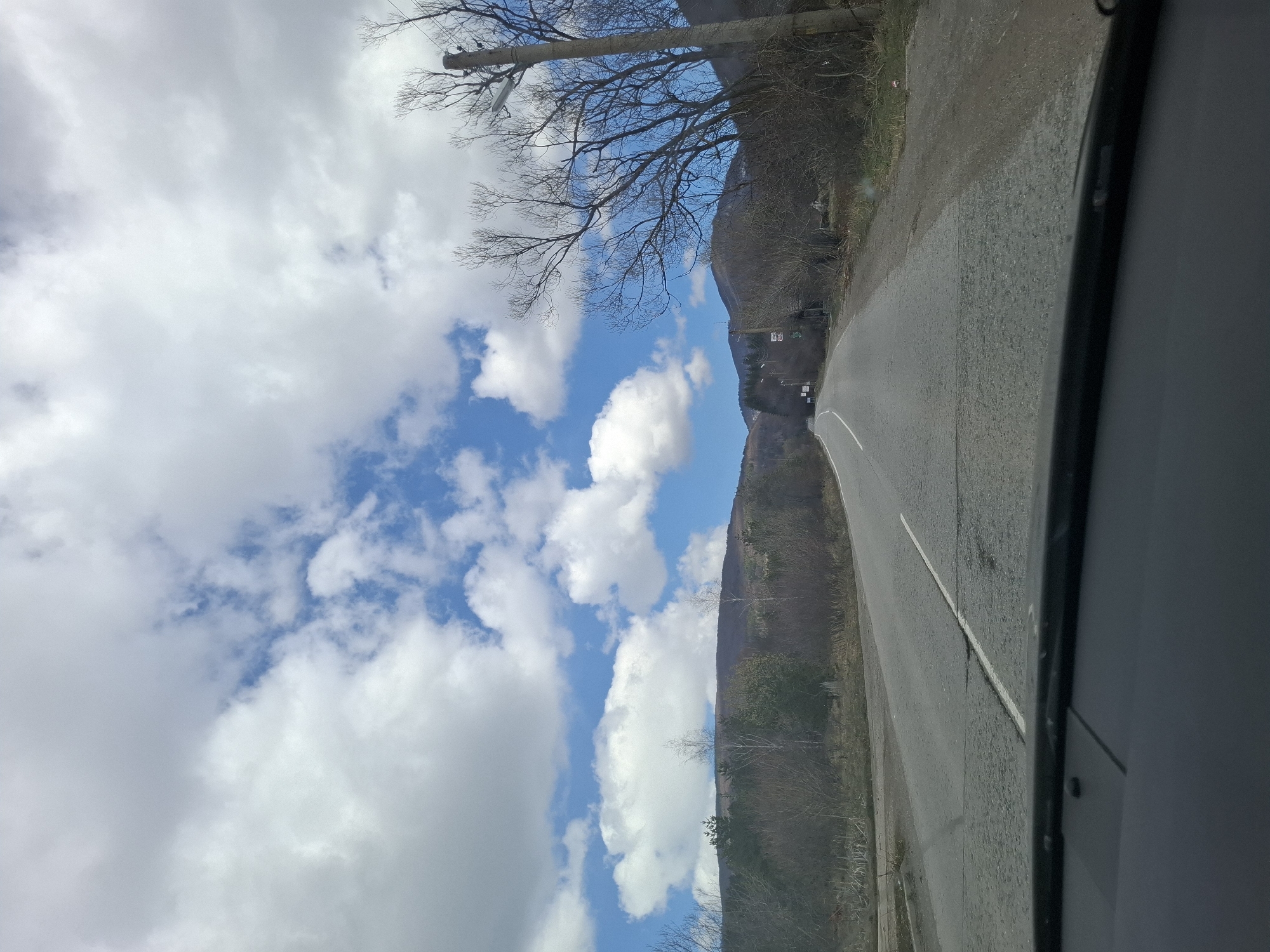 A view from inside a vehicle looking towards a paved road in Gintsi, Bulgaria. The road has a white dividing line. To the left of the road, there are barren trees and a cloudy, blue sky. To the right, there is a wooded hillside. The vehicle's interior trim is visible on the right side of the frame.