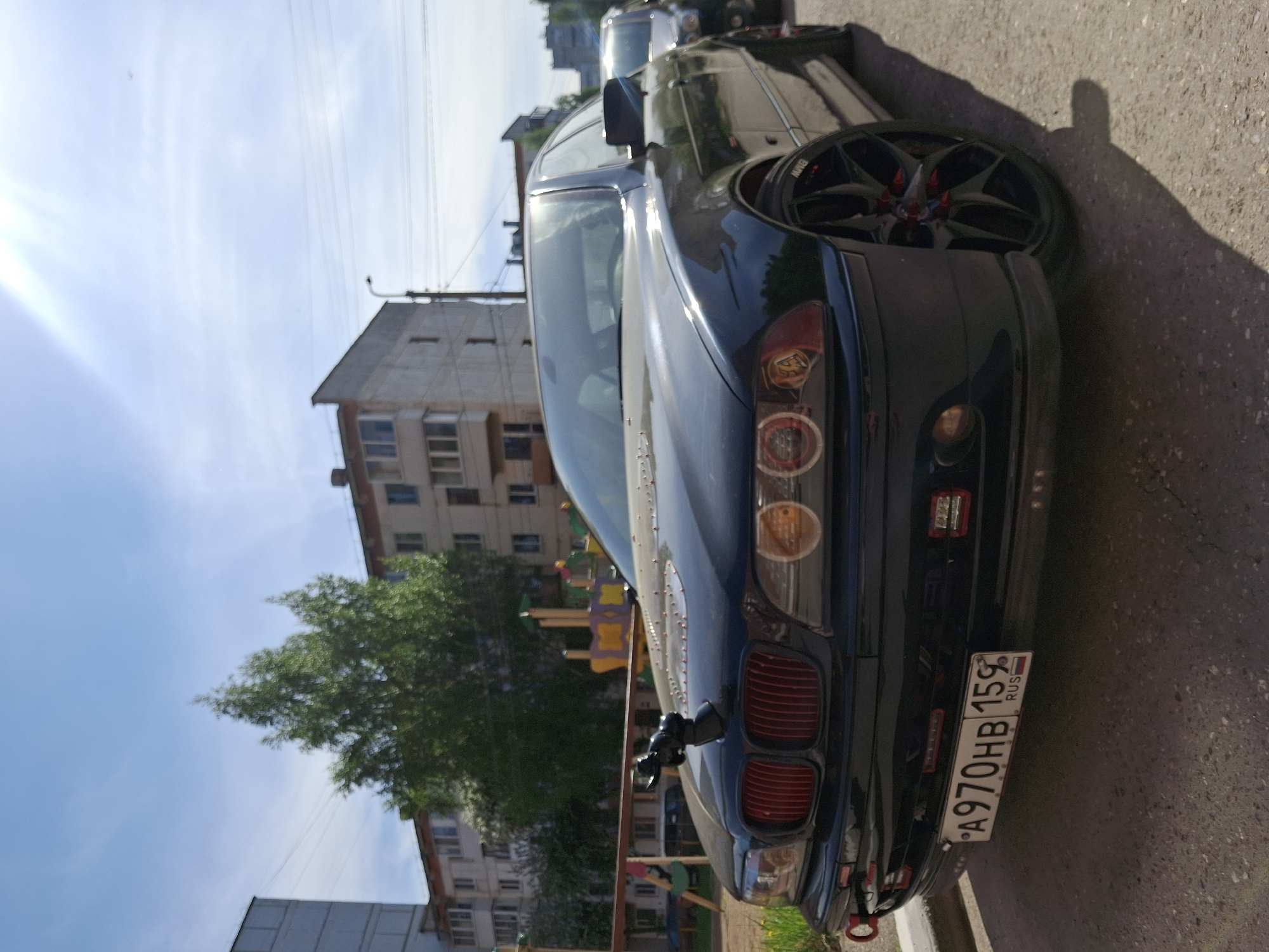 The image shows a dark-colored BMW car parked on a paved surface. The car's rear is prominently visible, including the license plate A970HB 159 RUS. The license plate indicates the car is registered in Russia's Perm Krai region (code 159). Buildings are visible in the background, along with a tree and a playground. Given the registration code and architectural style, the location is likely within the city of Chaikovsky, Russia, situated in Perm Krai.