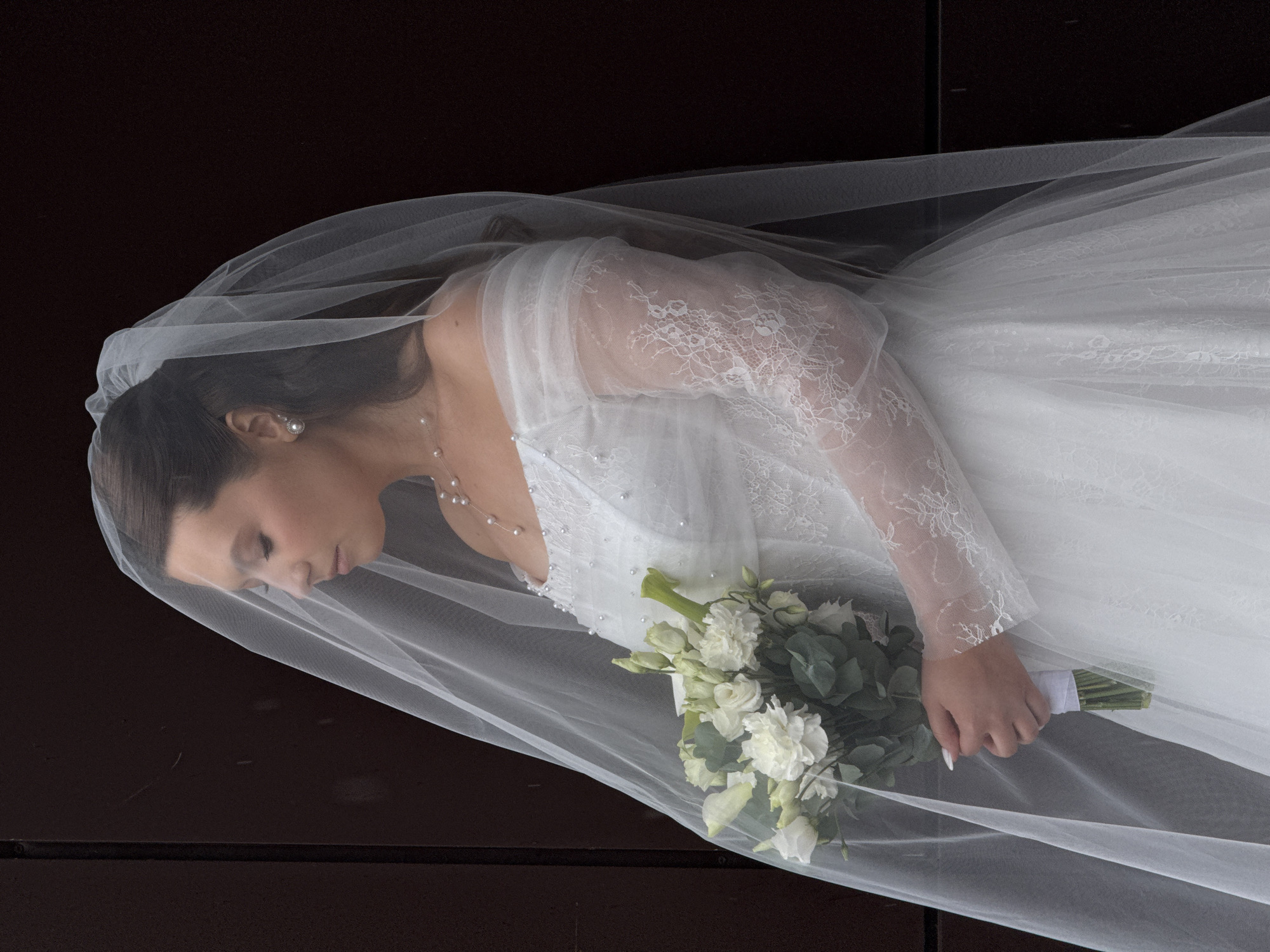A portrait-style photograph captures a young woman, presumably a bride, diagonally against a dark, featureless background. She is seen from the chest up, with her head tilted down and eyes closed or looking downwards, conveying a serene or contemplative expression. Her dark hair is mostly covered by a delicate, sheer white wedding veil that cascades around her head and shoulders, softly framing her face. She wears pearl stud earrings and a thin necklace with small pearls.
The bride is dressed in a white wedding gown featuring intricate lace long sleeves and a bodice with subtle embellishments, possibly beads or sequins. Her left hand, with manicured white nails, gently holds a white and green bouquet of flowers. The bouquet contains several white blossoms, possibly roses or similar types, interspersed with lush green foliage. The stems are wrapped in white ribbon.
The veil creates soft layers and textures throughout the image, partially obscuring parts of her dress and body in a delicate manner. The lighting is soft, highlighting the textures of the dress, lace, and veil against the stark, dark backdrop. There is no visible text or explicit location cues in the image; the background is a uniform dark brown or black. The overall impression is one of elegance and quiet grace.