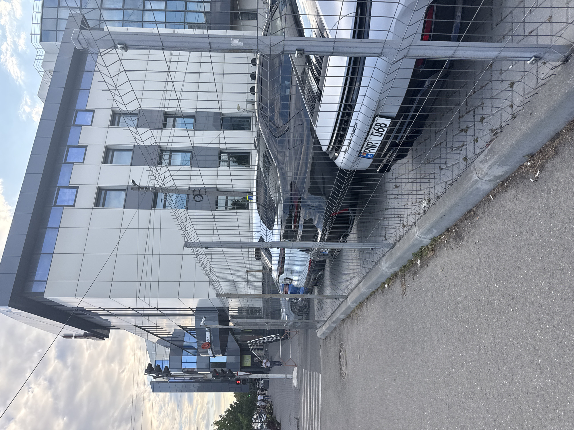 In the city of Chișinău, Moldova, a modern, multi-story building with a gray and white facade and blue-tinted windows stands behind a metal chain-link fence. The building has several floors with numerous windows. A sign for "SECURITY" is visible on the lower part of the building. In front of the building, parked vehicles are visible behind the fence, including a white car with a license plate that reads "PNR 068 MD". The area in front of the fence is a paved street with a sidewalk and a pedestrian crossing. Traffic lights are also visible in the background. The sky is cloudy.