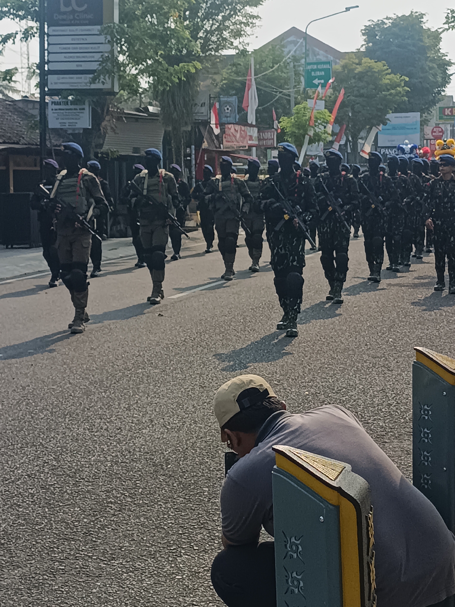 In Sleman, Indonesia, a procession of soldiers marches down a paved street. The soldiers are dressed in dark camouflage uniforms and blue berets, carrying rifles. They are walking in formation, with several rows of soldiers visible. In the foreground, a person wearing a light-colored cap and a gray shirt is crouching down, appearing to be taking a photograph or video with a phone. Behind the soldiers, buildings and trees line the street. Signs on a building indicate "Deeja Clinic" and "Praktek Dokter". Another sign points towards "Kantor Lurah Condongcatur" with an arrow. The scene appears to be a public event or parade.