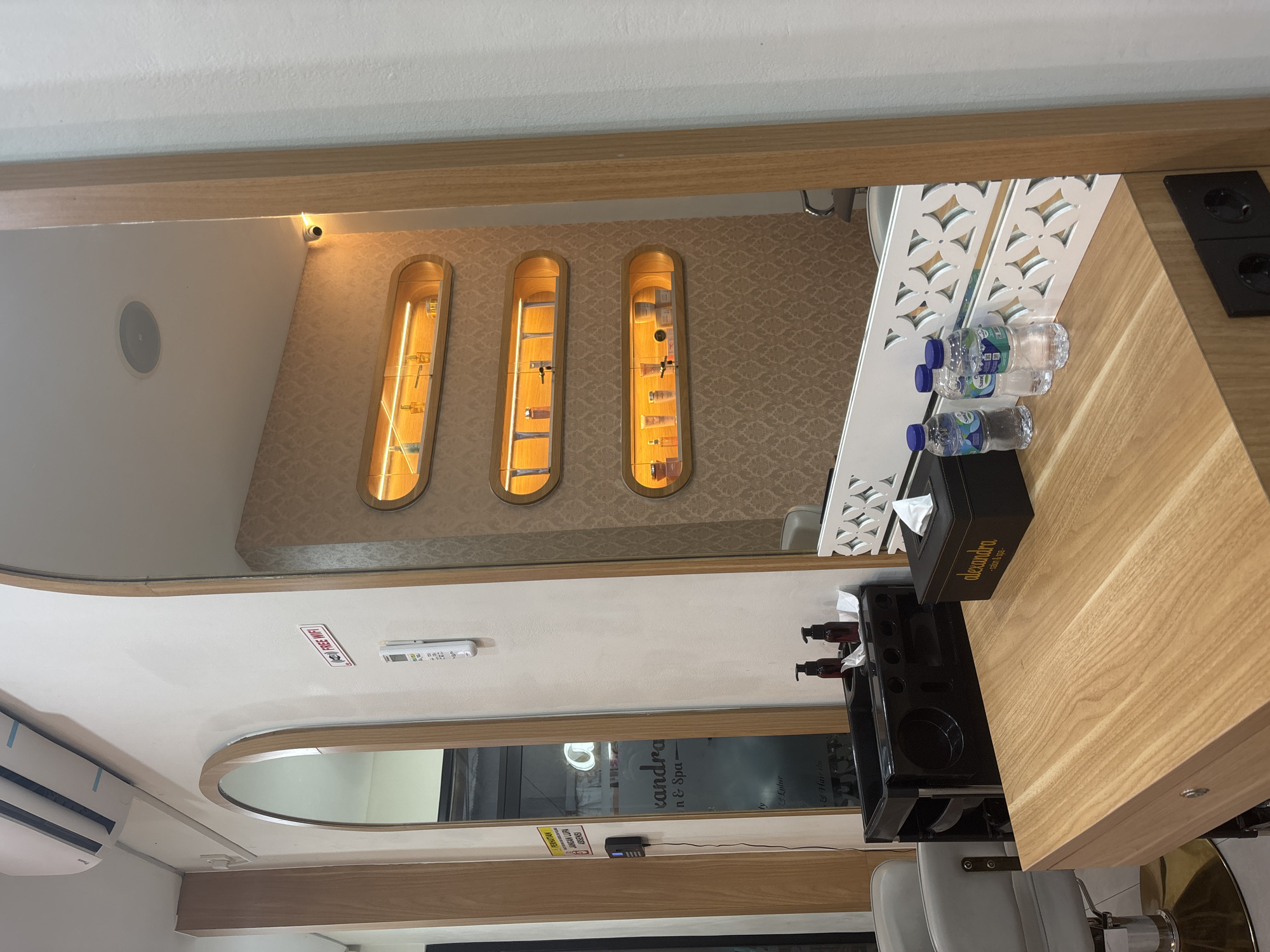 The image shows the interior of a business, specifically a salon or spa named "alexandra," as indicated on a tissue box and window. This salon likely offers services such as hair styling ("Hair do") and body treatments ("Lulur"). The presence of a "FREE WIFI" sign suggests an amenity for customers.
The layout includes mirrored surfaces, reflecting portions of the room and creating a sense of depth. There are built-in illuminated display niches with beauty products, and a workspace with several water bottles and a dispenser filled with products.
The environment appears well-lit, clean, and modern, with a focus on customer comfort, as suggested by the salon chairs. The location is in North Jakarta, Indonesia as indicated by signs.