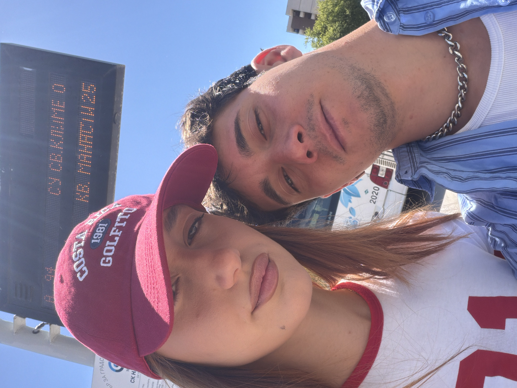 The image is a selfie of two individuals, a male and a female. The female is wearing a maroon baseball cap with white lettering that reads "COSTA RICA 1981 GOLFITO." She is also wearing a white T-shirt with a red collar. The male is wearing a light blue striped shirt over a white tank top and a silver chain.
In the background, there is a bus stop sign with an LED display that reads "СУ СВ.КЛИМЕ O KB. MAHACIN 25" in Cyrillic. This sign indicates the location is near the St. Kliment Ohridski University in Sofia, Bulgaria. Also, a bus or van with "2020" is partially visible behind the male.
