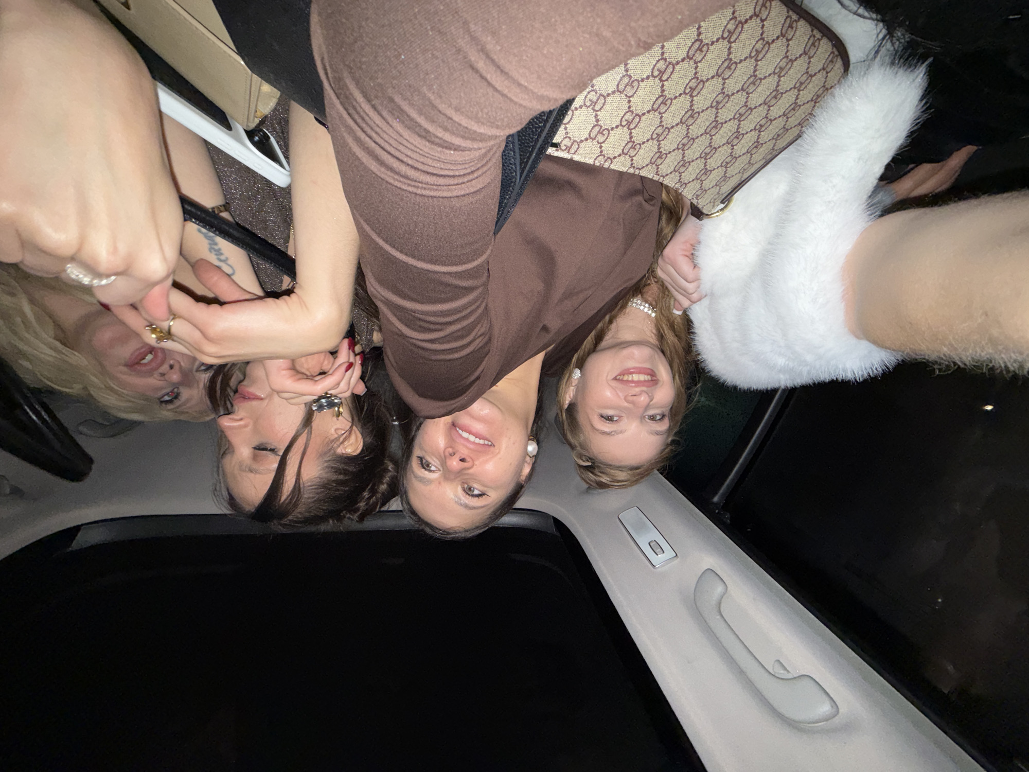 Four women are in the passenger compartment of a car. Three of the women are fair-skinned with light brown or blonde hair, and one has darker brown hair. One woman wears glasses, one wears a pearl necklace, and one has a tattoo on her inner left arm. One woman holds a white furry object. A brown purse with a repeating pattern is near one woman. The car has a sunroof, and there is a hand grip on the interior roof. It is dark outside. The location is unknown.