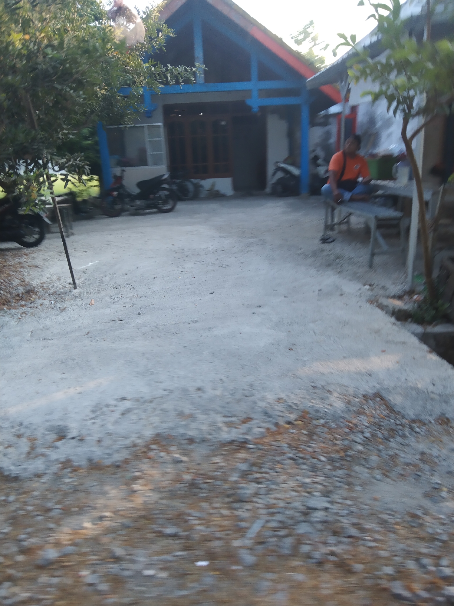 The image depicts an outdoor scene in the city of Growok, Indonesia, likely a residential area. In the foreground, a rough, gray, gravelly surface dominates, with some scattered stones and dry leaves. A thin, dark, vertical pole is planted in this surface on the left side.
In the middle ground, a paved area transitions to the gravel. A man wearing an orange shirt and blue pants is seated on a gray, slatted bench to the right. He has a dark bag slung over his shoulder and appears to be holding something in his hands, possibly a phone. Next to him, a small tree with green leaves grows.
In the background, a two-story building with a blue-trimmed, red-roofed upper section is visible. The lower section of the building is white with brown-framed windows and doors. Two motorcycles are parked in front of the building, one partially obscured by a tree on the left. Lush green trees are present on the left side of the image, with dense foliage. The overall impression is a somewhat rustic, lived-in environment.