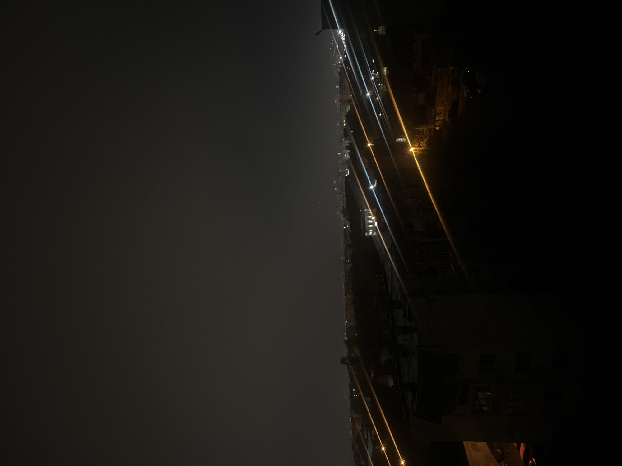The image displays a nighttime urban landscape in Sofia, Bulgaria, viewed from a slightly elevated perspective. The dominant feature is a vast, dark, and seemingly misty or overcast sky, occupying the upper left portion of the frame (when correctly oriented). This dark expanse transitions to a line of artificial lights that stretch horizontally across the middle of the image, depicting a city or street below.
These lights are a mix of bright white/blue and warm yellow/orange, appearing as distinct, elongated streaks, possibly due to a long exposure photograph or the diffusion of light through atmospheric moisture like fog or mist. Below and to the right of these light streaks, indistinct dark silhouettes of buildings and some sparse vegetation can be seen, partially illuminated by the city's glow. The scene suggests a quiet or distant view of an urban area after dark. No people or specific activities are discernable, only the ambient illumination of the city. The time of day is clearly night, and the atmospheric conditions appear to be cloudy or foggy, enhancing the streaking effect of the lights. No text is visible in the image.