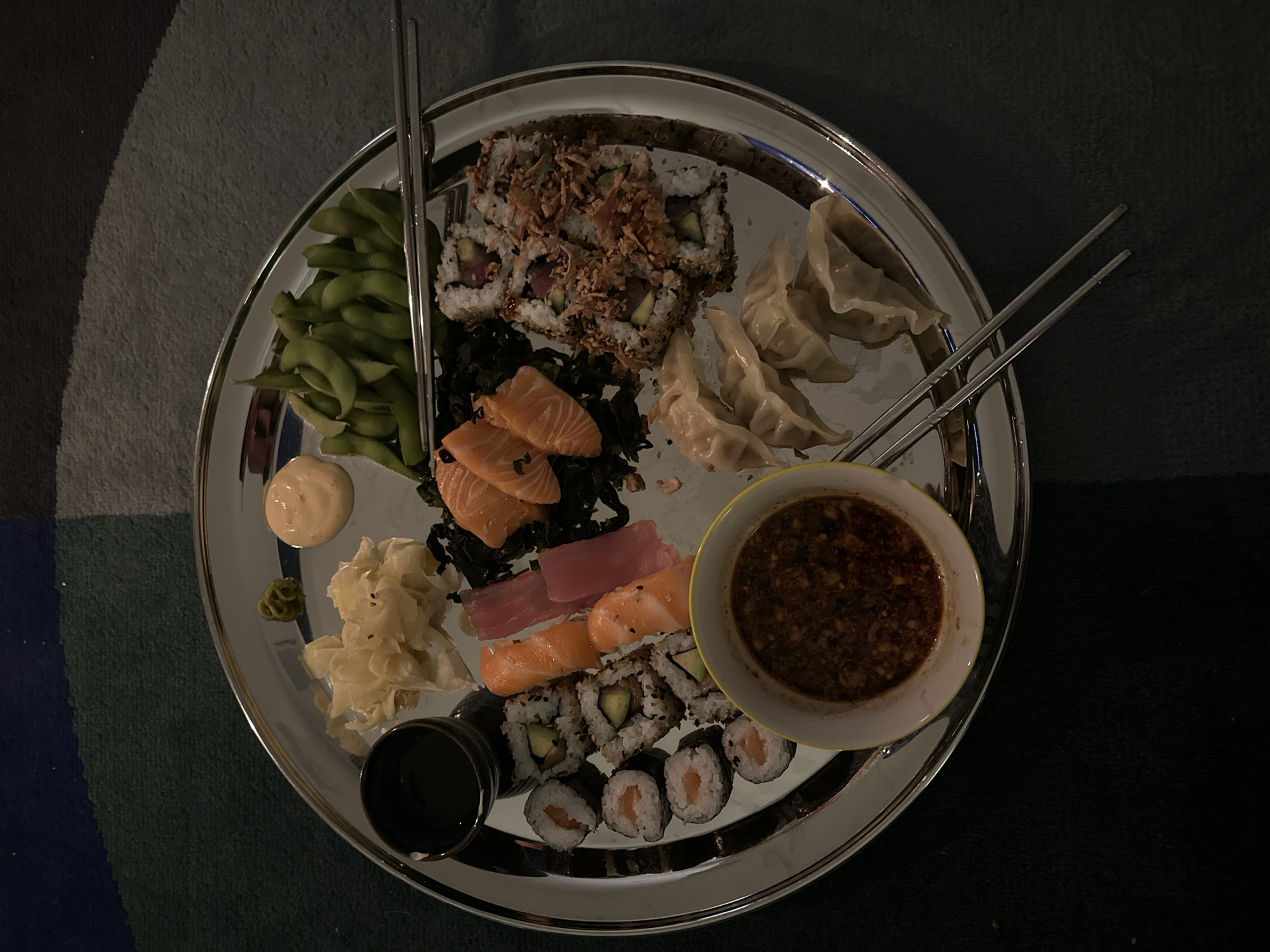 A silver platter of sushi and Asian food is centered in the image. The platter includes edamame, dumplings, various sushi rolls, slices of salmon and tuna, ginger, wasabi, two sauces, and chopsticks. The platter rests on a rug with blue, green, gray and black sections. The lighting is subdued. The location is not identifiable as being within London or the United Kingdom. No people are visible in the image.