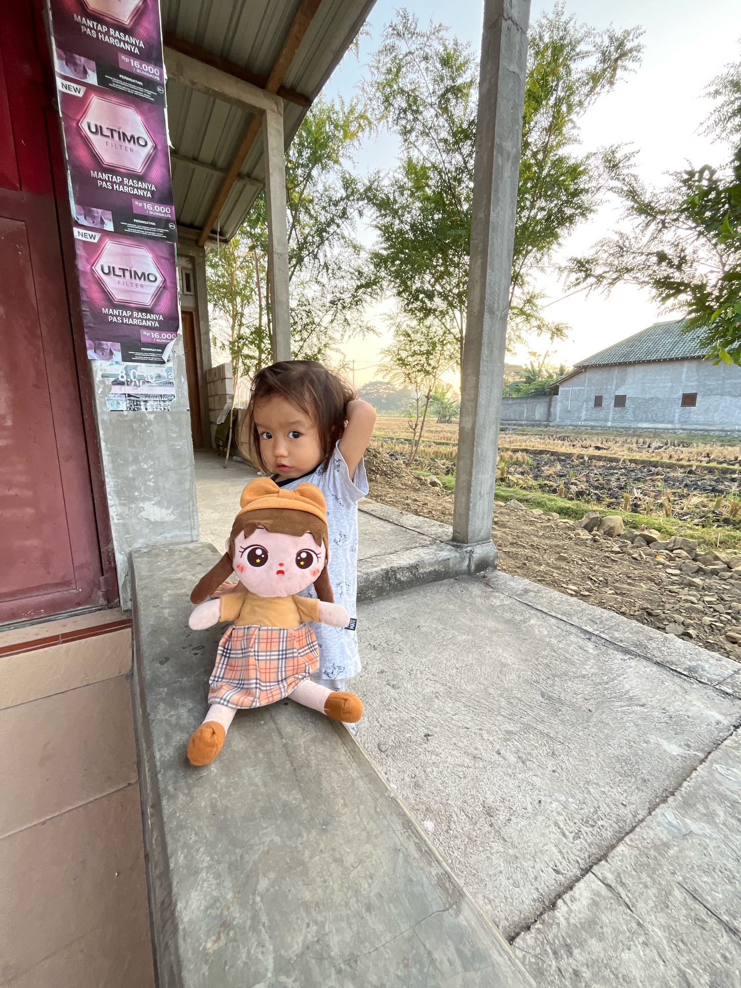 In Kauman, Indonesia, a young child stands beside a plush doll on a concrete ledge. The child, with dark hair and fair skin, wears a light-colored t-shirt and appears to be touching their hair. The doll, dressed in a plaid skirt and orange top, is positioned in front of the child. Behind them, a building with a corrugated metal roof is visible, along with a field containing agricultural residue. The scene is lit by natural light, suggesting it is daytime. Posters are affixed to the wall near the building entrance, and a concrete pillar stands to the right of the child.