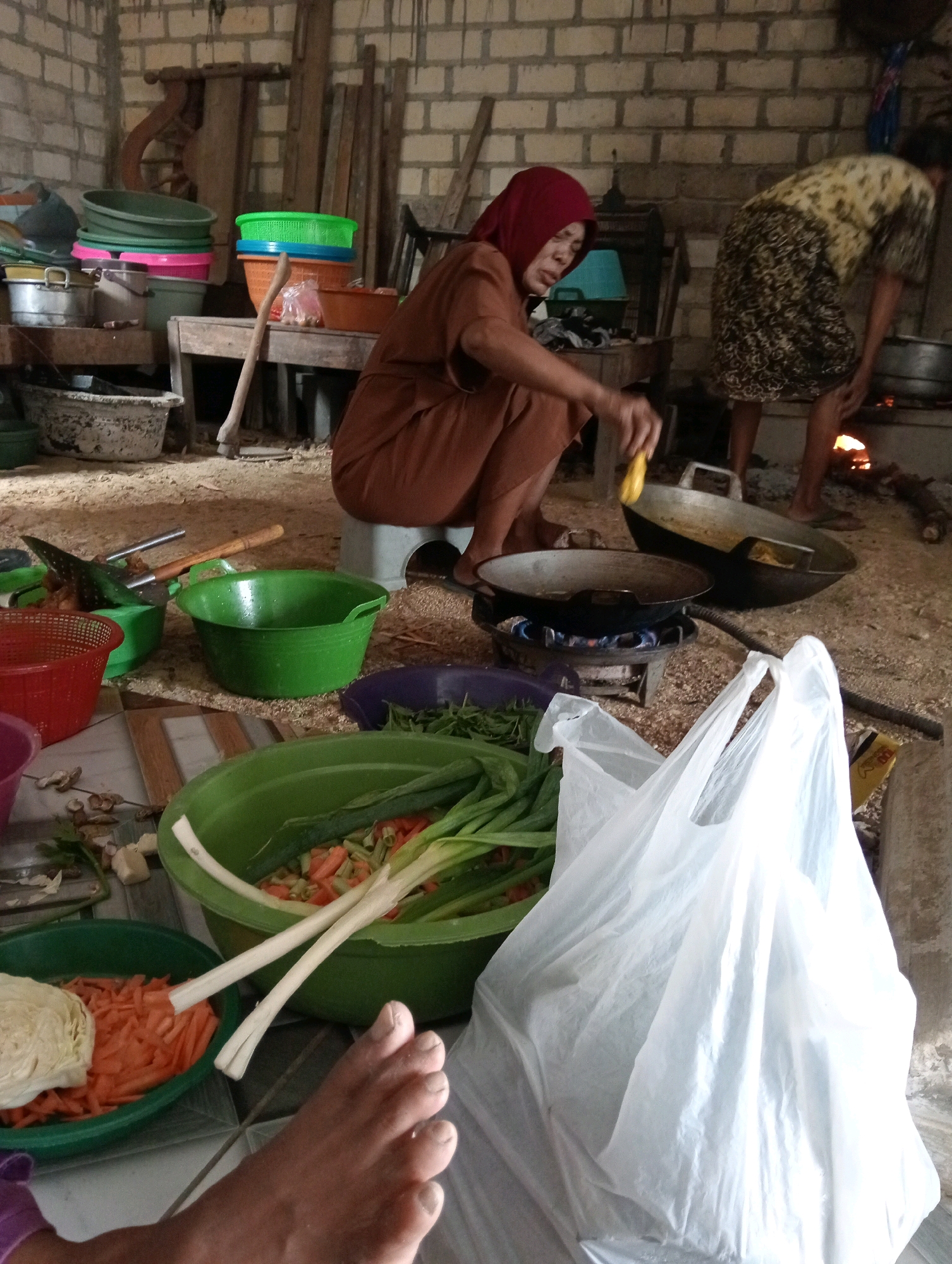 The image depicts an outdoor kitchen scene in Soko, Indonesia. Two women are present. One woman, wearing a red headscarf and a brown outfit, is crouching and stirring food in a wok over a small stove. The second woman, in a patterned dress, is bent over and attending to a larger cooking vessel on a fire pit in the background. Various cooking implements, including woks, bowls, and baskets, are scattered around the area, along with vegetables such as carrots and green onions. The ground is covered in dirt and wood shavings. The environment appears rustic, with a brick wall in the background and wooden structures. A white plastic bag is prominently displayed in the foreground.