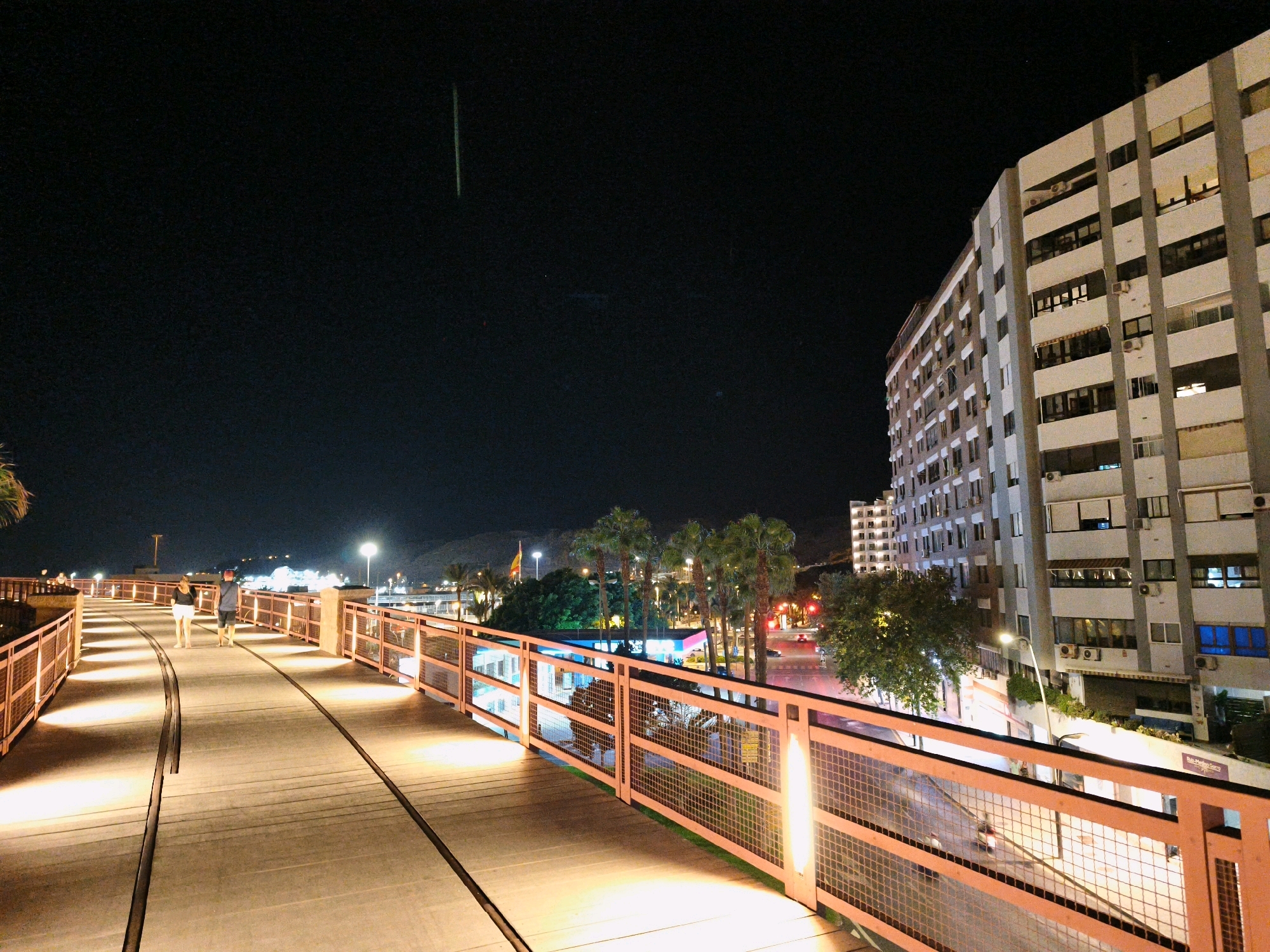 The image depicts a pedestrian bridge illuminated by warm lights at night in Almeria, Spain. The bridge has a wooden deck with two sets of dark tracks running parallel down its length. The bridge is bordered by orange metal railings with mesh infill. Two individuals, a man and a woman, are walking away from the camera on the bridge. In the background, a large, modern, multi-story building with many windows stands prominently. Palm trees and other vegetation are visible, along with streetlights and the lights of businesses. A faint streak of light, possibly a meteor or satellite, is visible in the dark night sky. The overall scene suggests an urban environment at night with a focus on the illuminated pedestrian walkway and the surrounding cityscape.