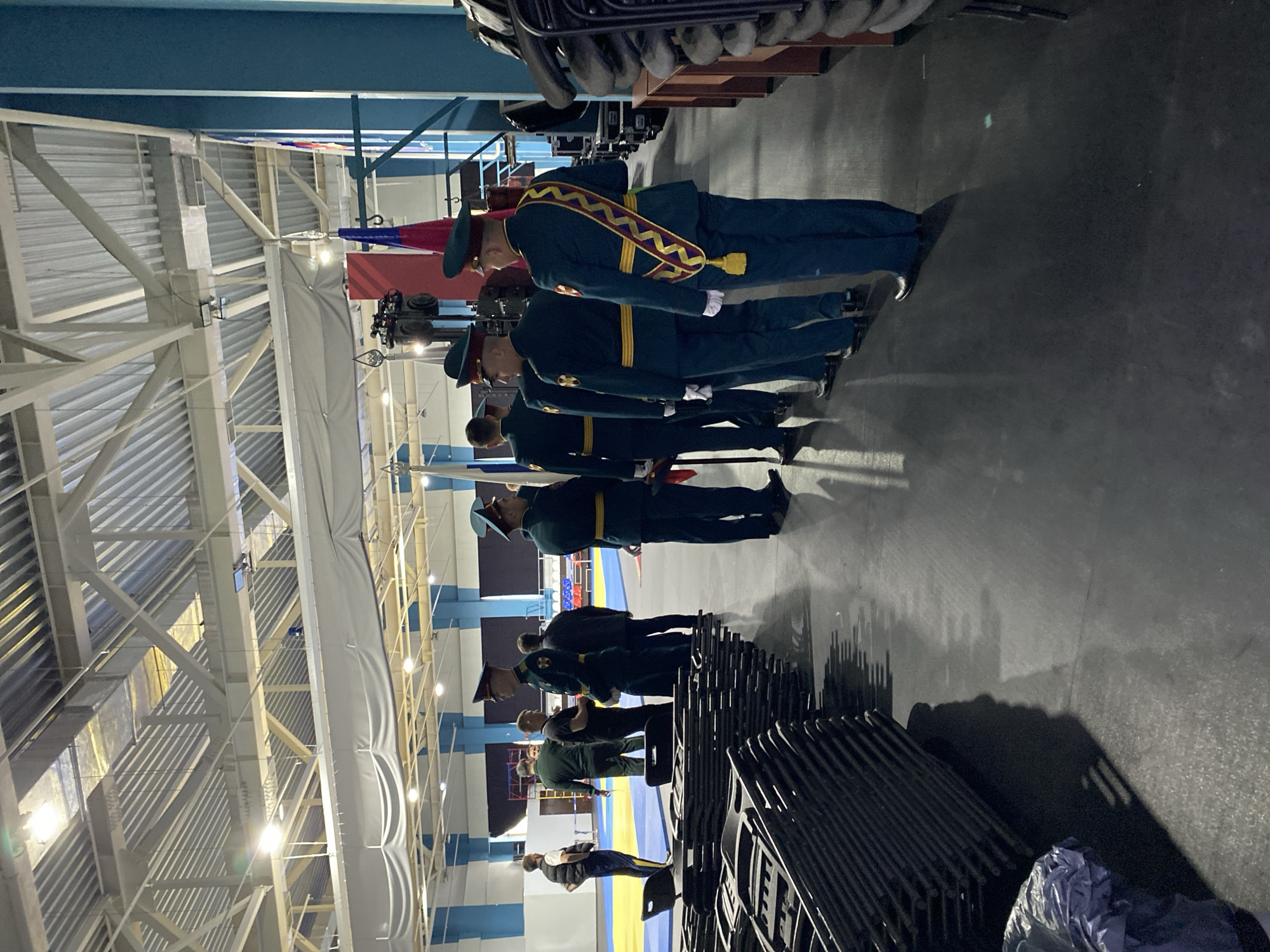 The image captures an indoor scene, likely a large hall, gymnasium, or convention center, characterized by a high ceiling with visible girders, trusses, and corrugated metal panels, illuminated by artificial lights. A long, grey fabric banner or screen hangs vertically along one wall.
The central subjects are a group of men, predominantly those in military-style uniforms. A line of men in dark green uniforms, featuring gold trim and peaked caps, stands formally. The man at the front of this line is distinct with a prominent yellow and red patterned sash worn diagonally across his chest. Further back, more individuals are visible; some also wear uniforms, while others are in civilian clothing. One person in the background wears a white shirt and dark vest.
The activity appears to be a formal gathering, ceremony, or inspection, given the disciplined formation of the uniformed personnel. The mood conveyed is serious and formal.
Notable objects include large stacks of dark-colored chairs in the foreground and on the right side, suggesting seating for an event or audience. In the background, on the floor, a prominent blue and yellow mat or banner is visible. No discernible text is visible anywhere in the image. The lighting is artificial, casting various shadows across the floor and walls.