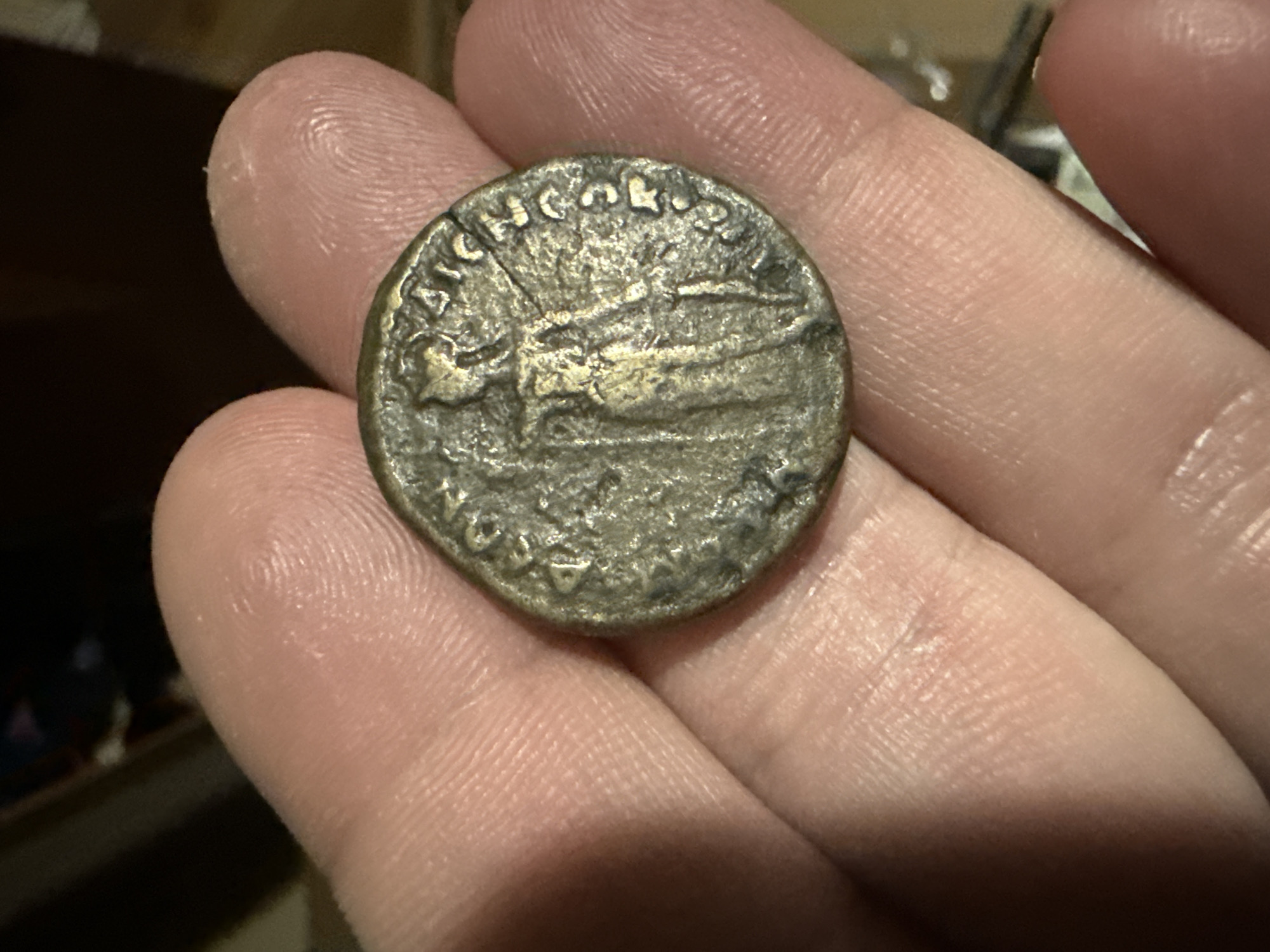 A close-up image captures a person's hand holding a single, dark-colored coin. The hand is positioned so that the coin rests on the fingertips and palm, allowing for a clear view of its obverse (or reverse, if the other side is less detailed). The skin of the hand shows visible fingerprints and fine lines, indicating artificial or low light conditions with shadows.
The coin itself appears to be an ancient metallic artifact, likely made of bronze or a similar copper alloy, given its dark patina and hints of green oxidation. It has an irregular, somewhat worn circular shape. On the visible side of the coin, there is a distinct effigy of what appears to be a figure, possibly a Roman emperor or deity, facing to the right on the left side of the coin. To the right of this figure, a larger, elongated object is depicted, which strongly resembles a galley or ship, indicating a possible naval theme.
Around the upper edge of the coin, part of an inscription in capital letters is visible, though somewhat worn. Recognizable letters include "LIC" and "CON," possibly part of an emperor's name such as Licinius, which was common on Roman coinage. Further letters and symbols are partially visible along the lower edge, but are less distinct.
The activity shown is the examination or presentation of this historical coin. There are no direct cues within the image to confirm the provided location context of Krumovgrad, Bulgaria, as the background is out of focus and nondescript, focusing solely on the hand and the coin.