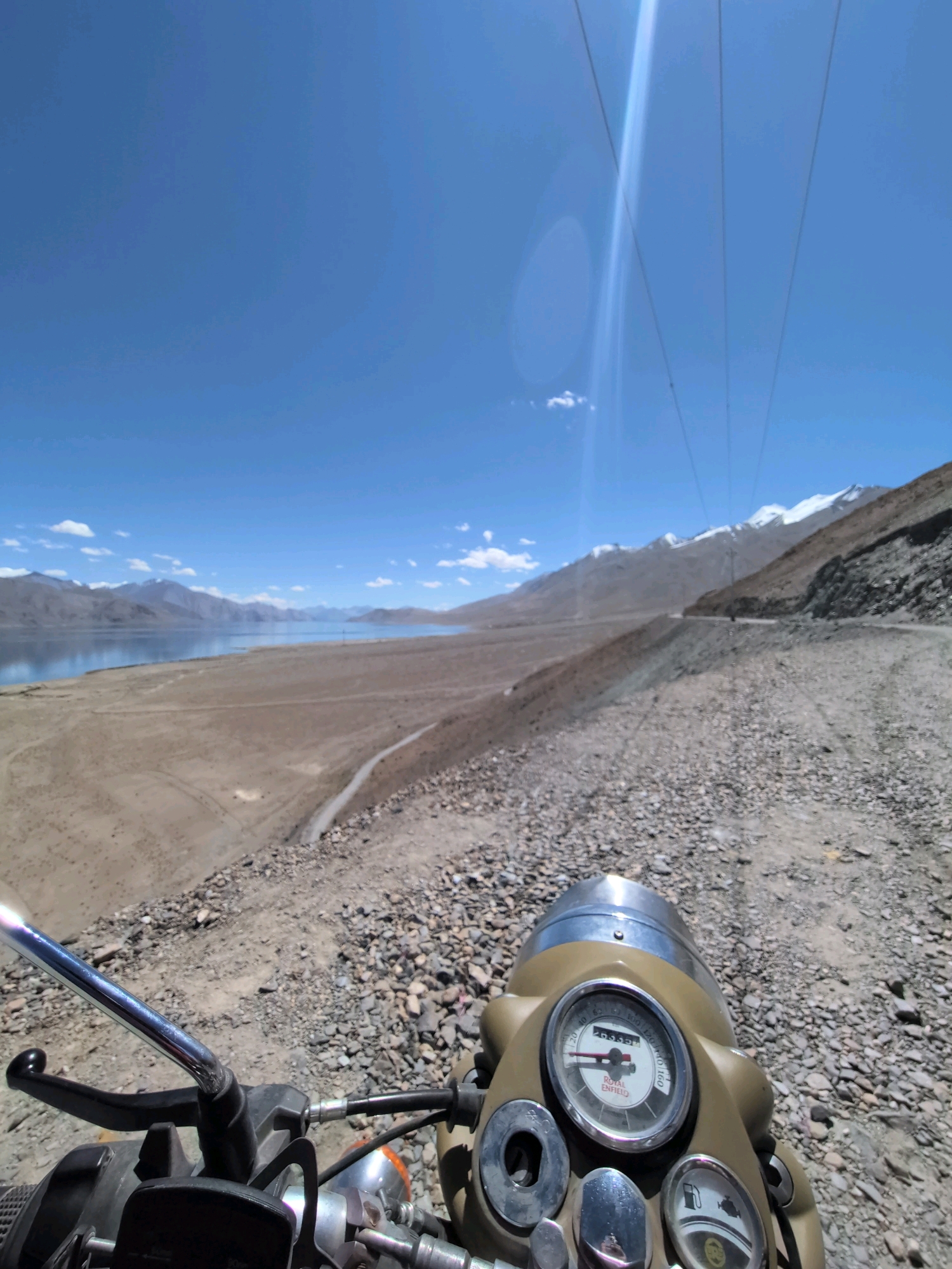 A first-person perspective shot from a motorcycle shows a scenic landscape under a clear blue sky. The foreground is dominated by the motorcycle's handlebars, mirrors, and instrument cluster, including a speedometer and a fuel gauge. The road ahead appears to be unpaved and rocky, winding through a barren, sandy terrain. In the distance, a large, calm body of water stretches out, reflecting the sky and surrounded by rugged, mountainous terrain. The mountains in the background are imposing, with some peaks possibly snow-capped. Power lines are visible stretching across the sky, supported by poles. The overall impression is one of remote, natural beauty, possibly in a high-altitude desert region of India.
