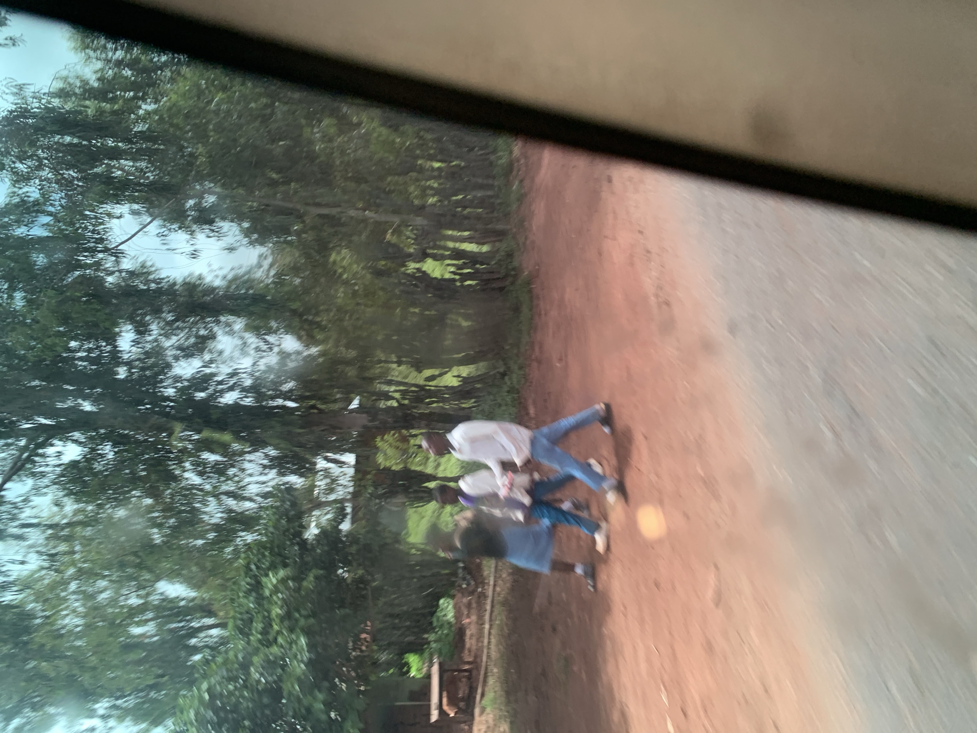 Three young people are walking along a reddish dirt road. The person on the left is wearing a dark shirt and a blue skirt. The person in the middle is wearing a white shirt with purple accents and blue pants. The person on the right is wearing a white long-sleeved shirt and blue pants. They are walking away from the viewer. In the background, there are tall trees and some low buildings. The scene appears to be outdoors, possibly in a rural or semi-rural area of Hoss, Nigeria, given the unpaved road and vegetation.
