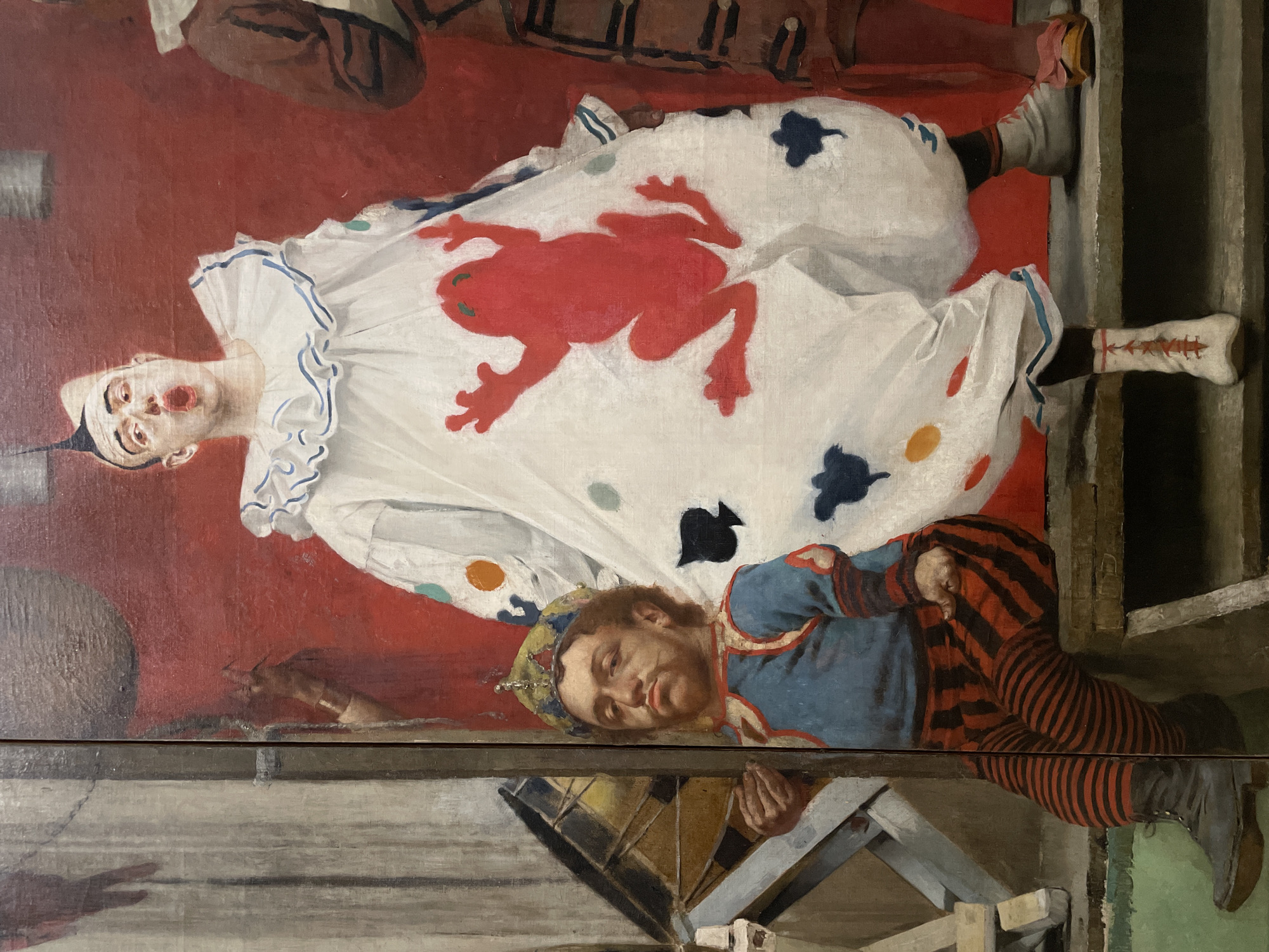 This image is a photograph of "The Clowns" (also known as "The Harlequin and the Dwarf"), an oil painting by Edgar Degas, located in the Musée d'Orsay in Paris, France.
The painting depicts two primary figures in theatrical costumes against a predominantly red backdrop, suggestive of a stage curtain. On the right, a tall figure in a voluminous white clown suit with a ruffled collar stands with an open mouth, as if shouting or singing. This figure has white face paint, a spiky dark coiffure, and is adorned with a large red frog silhouette on the torso, along with smaller orange, teal, and black irregular shapes across the costume. White laced shoes are visible on their feet. To the left, a shorter figure, a man with a reddish-brown beard and mustache, sits on a wooden platform. He wears a blue top with red trim, red and black striped leggings, and black boots, topped with a golden, slightly dented crown. His expression is somber, and he looks directly forward, holding a small, indeterminate object in his right hand. To the far left, partially obscured, is a drum on a stand, and behind the seated figure, a wooden structure is visible. Another figure, largely out of frame to the far right, is partially visible, wearing a brown costume. The figures are positioned on a multi-tiered wooden stage or platform.