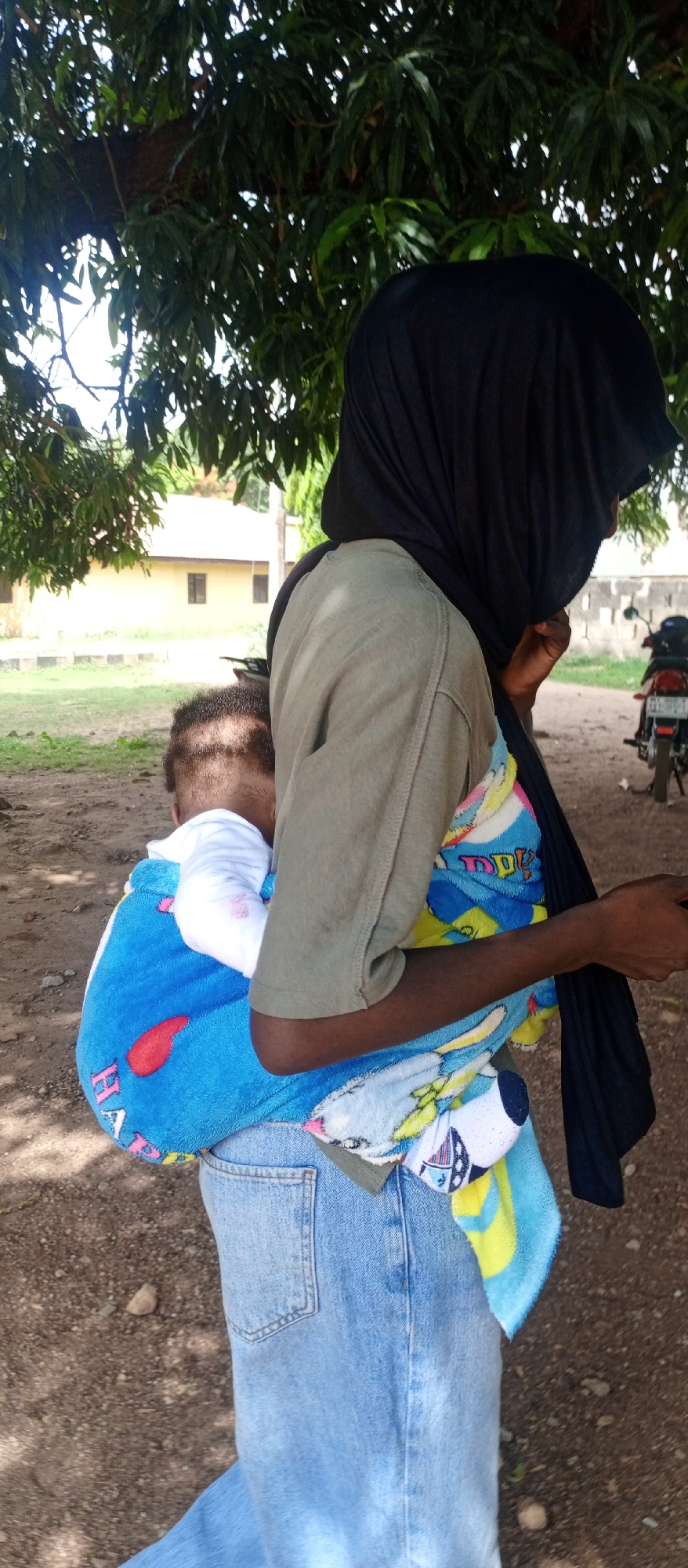 A woman in Keffi, Nigeria, is seen from the side carrying a baby in a sling on her back. She wears a black hijab, a khaki-colored shirt, and blue jeans. The sling is made from a blue and multi-colored patterned towel. A portion of the baby's head and arm are visible. The scene is outdoors, under the shade of a tree, with a building and a motorcycle visible in the background. The ground appears to be dirt or gravel.