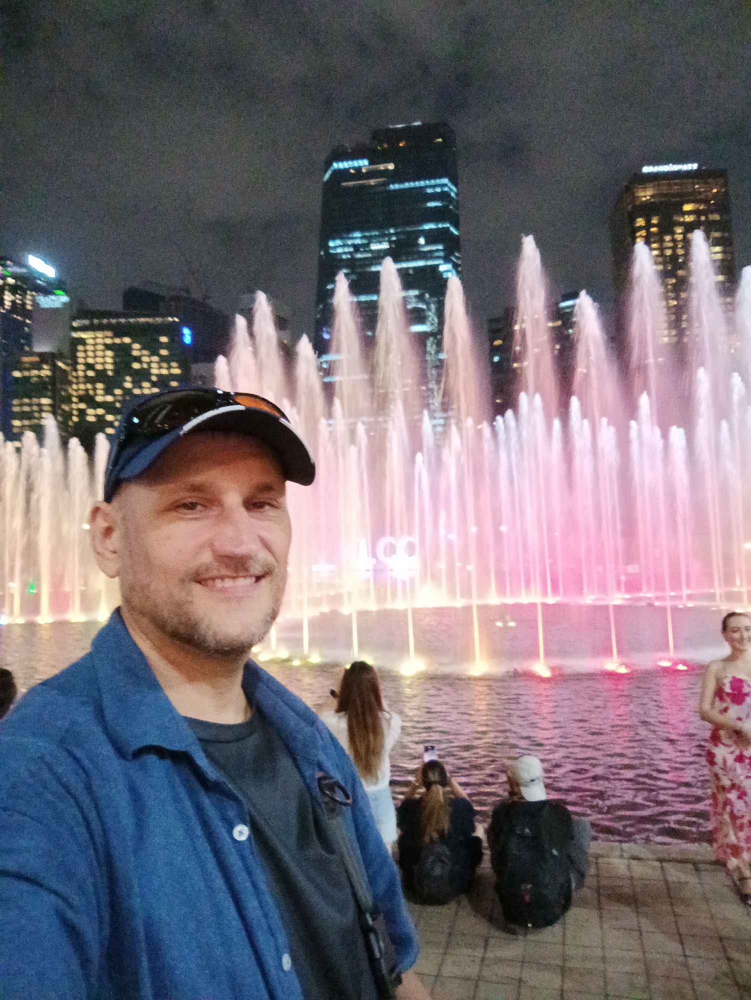 A medium shot captures a man in the foreground, wearing a blue jacket over a dark shirt and a blue baseball cap with sunglasses perched on top. He is smiling directly at the camera. Behind him, a vibrant fountain display illuminates the night sky with streams of pink and white water. In the background, towering city skyscrapers with illuminated windows punctuate the darkness. To the right, a woman in a floral dress stands near the fountain. Other individuals are partially visible in the middle and background, some seated and others standing, observing the fountain. The scene appears to be set at the KLCC Park in Kuala Lumpur, Malaysia, judging by the prominent Petronas Twin Towers and the illuminated cityscape characteristic of the area.
