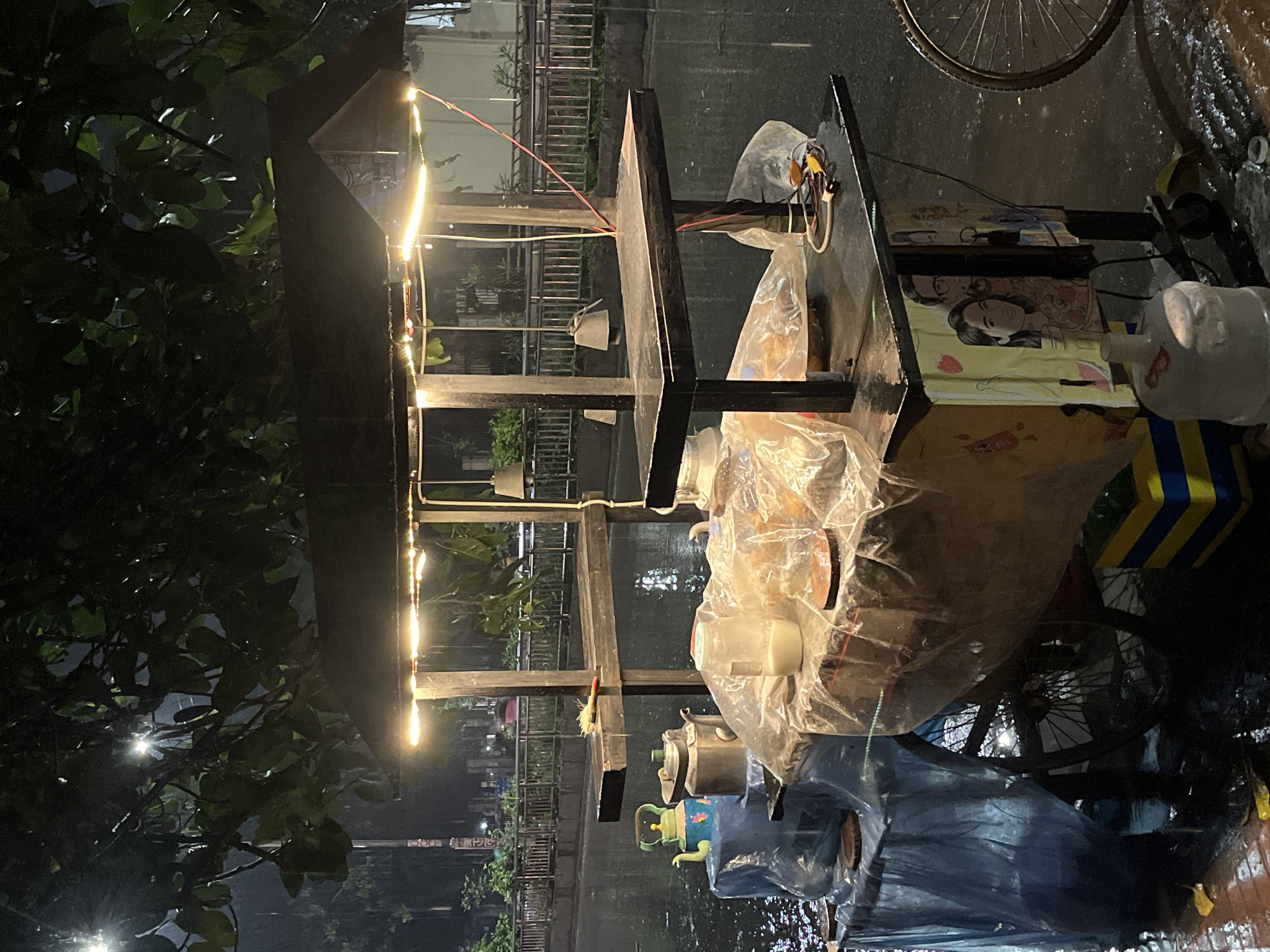 The image shows a street food cart in Dhaka, Bangladesh at night during rainy weather. The cart is covered in plastic sheeting and lit by a string of lights. Various pots, containers, and food items are visible on the cart. The surrounding environment includes trees, a fence, and a wet street.