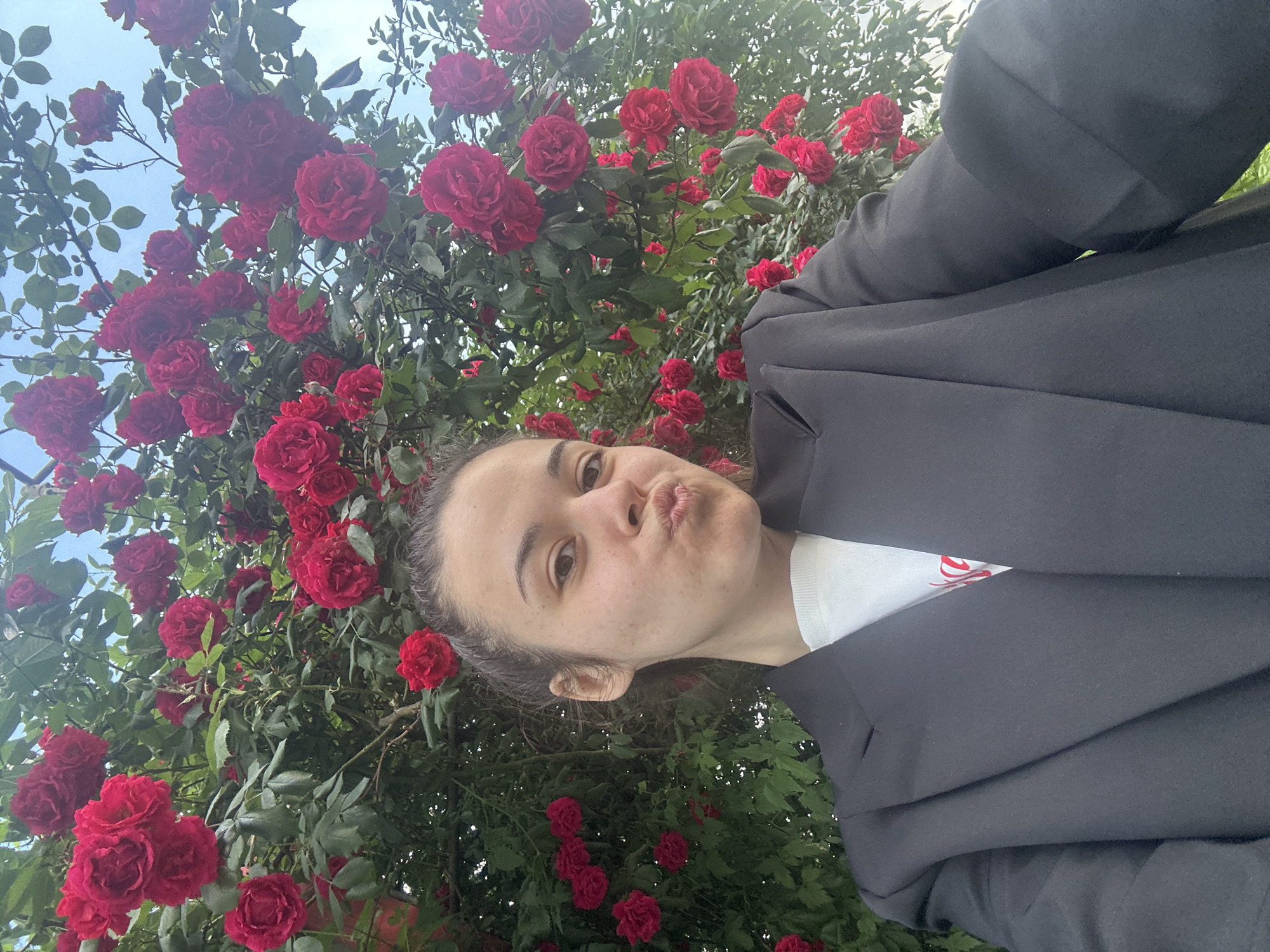 A close-up selfie captures a woman looking up towards the camera with puckered lips, indicating a playful expression. Her head and upper body are visible, framed by a dense and vibrant backdrop of numerous red roses and green leaves. Patches of clear blue sky are visible through the foliage, suggesting a bright, daytime setting under clear weather. The woman has her hair pulled back and visible freckles on her face. She is wearing a black blazer or jacket over a white top, on which indistinct red text or a logo is partially visible. The scene takes place outdoors, amidst what appears to be a large rose bush, possibly in a garden or park in Sofia, Bulgaria.