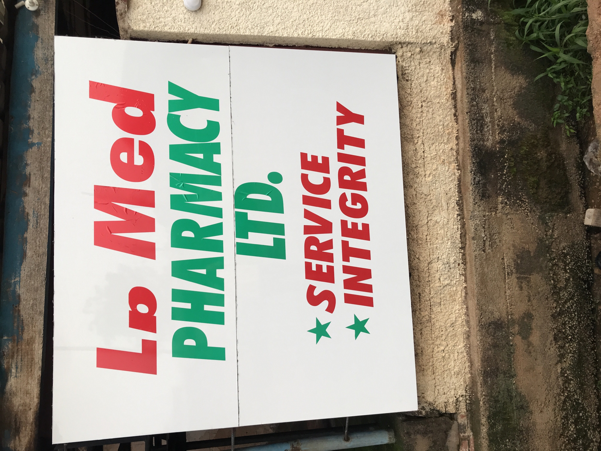 A white sign with red and green lettering is mounted on a rough, beige-colored wall, above a darker, textured surface. The sign is divided into two panels. The top panel displays "La Med PHARMACY LTD." in red and green. The bottom panel shows two green stars followed by "SERVICE" and "INTEGRITY" in red. The sign is located in New Stadium, Nigeria. The surrounding environment includes a rusty metal fence and some vegetation.