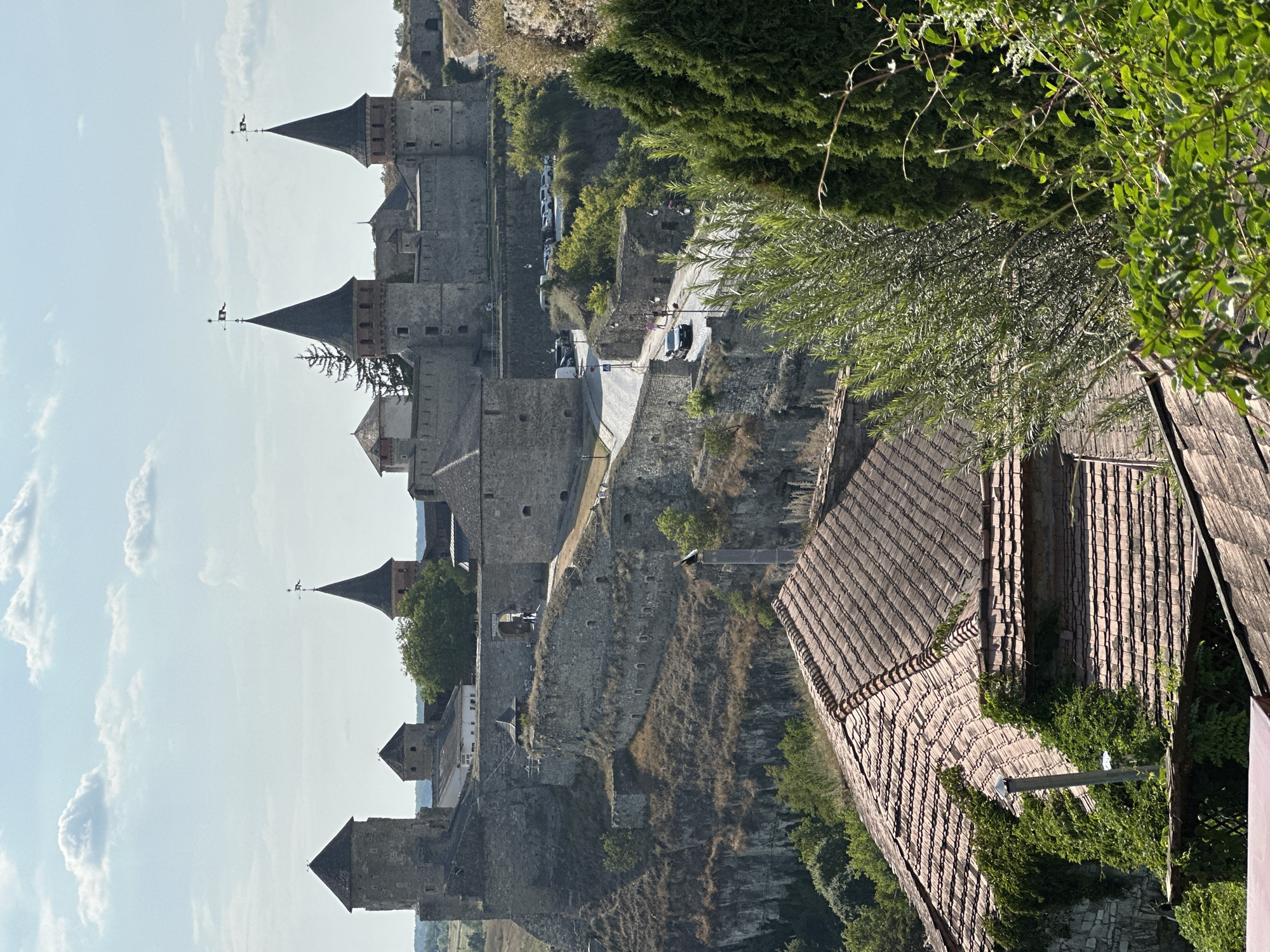This is a medium-distance, slightly elevated, eye-level shot of the Kamianets-Podilskyi Castle in Ukraine on a bright, partly cloudy day. In the foreground, the tiled roof of a building is visible, covered in ivy. Beyond this, a road winds its way up a hill towards the castle, with several cars parked on the side. The castle itself is a large, stone fortress with multiple towers, some of which have conical roofs topped with flags. The castle is situated on a rocky cliff face, with green trees and bushes growing on its slopes. The sky is a pale blue with scattered white clouds.