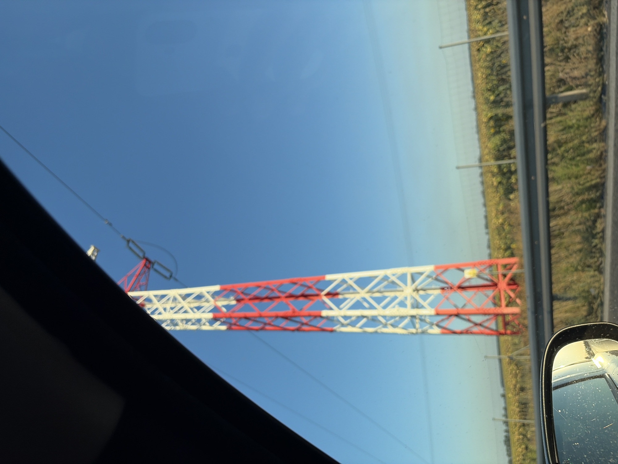 The image was taken from inside a vehicle, looking out through the windshield and side window. A tall, red and white lattice tower, likely a power pylon, is the central subject of the image. It extends high into a clear blue sky. The tower is partially obscured by the dark frame of the vehicle's interior. In the foreground, a metal guardrail runs horizontally across the scene, separating the road from a field of sunflowers that stretches towards the horizon. A fence can be seen behind the guardrail, and beyond that, the field of sunflowers with more distant trees or structures. The reflection in the side mirror shows the road and the sky, indicating the vehicle is in motion. The scene appears to be set in a rural or semi-rural area, possibly on the outskirts of Dolná Streda, Slovakia, given the sunflower fields and power infrastructure common in such landscapes.