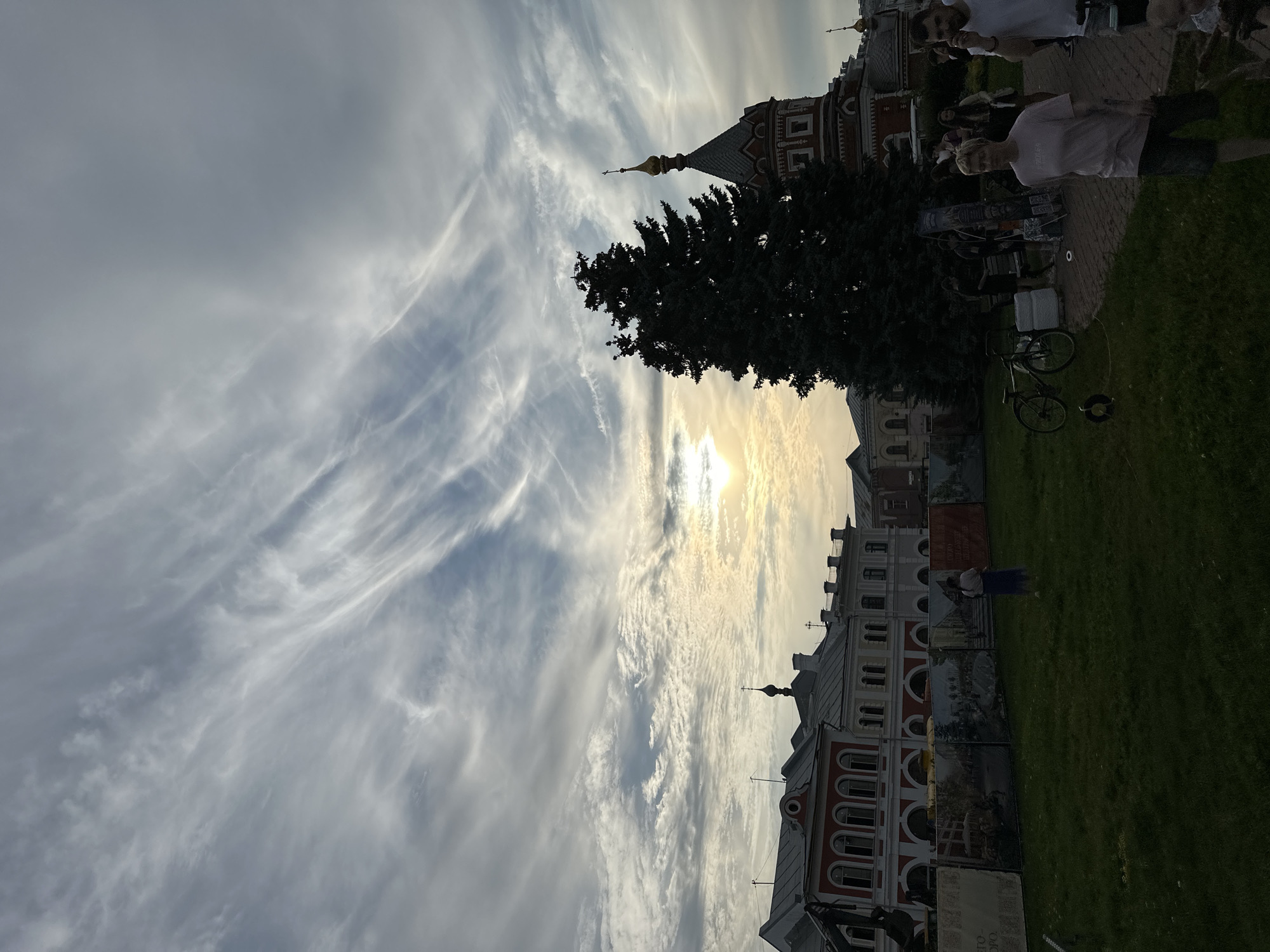 This is a medium-shot, eye-level, outdoor photograph taken in the city of Putyatino (Semivragovskaya S/A), Russia. The scene captures a broad view of a public space with several buildings, a large evergreen tree, and a portion of the sky.
In the background, a long, multi-story building with red and white facades is visible. The architecture features arched windows and decorative elements. To the right of this building, a portion of a church or religious building with a red brick lower level and a domed, golden-topped roof is present. The sky above is dramatic, filled with wispy, elongated clouds that stretch across the frame. The sun is setting or rising, creating a bright, hazy glow towards the upper right quadrant of the image.
In the mid-ground, a tall, dark green evergreen tree stands prominently, silhouetted against the sky and the buildings. In front of the buildings, there is a grassy area. A fence or barrier with large printed banners depicting various scenes, possibly historical or artistic, runs along the edge of the grassy area.
In the foreground and mid-ground, there are several people. On the far left, a person is standing on the grass facing away from the camera, wearing a light-colored top and a blue skirt. Towards the right, a group of people are walking on a paved path or area. One woman in the foreground is wearing a pink t-shirt and dark shorts, walking towards the camera. Another man in a white t-shirt is visible to her right, also walking. Other individuals are present in the background, some appearing to be interacting or browsing displays. A bicycle is parked on the grass to the left of the paved area. Several stacks of what appear to be white crates are also visible near the bicycle.
The overall atmosphere is that of a public gathering or event, possibly a park or historical site, during a time of transition in the day, indicated by the sky and sunlight.