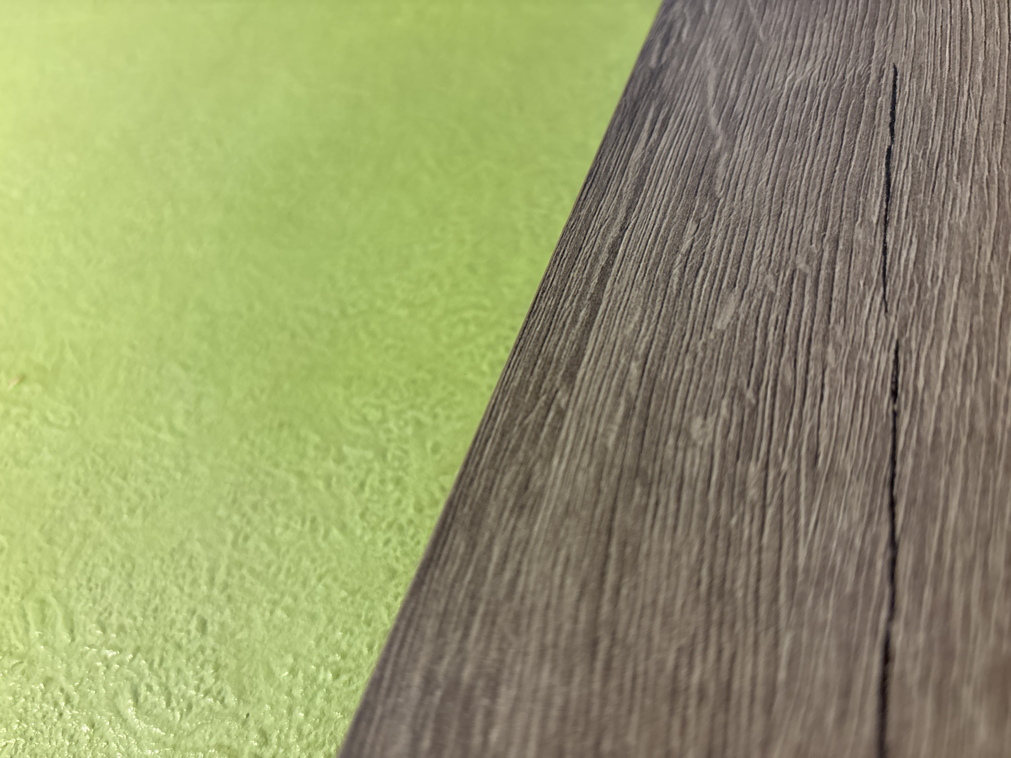 The image shows a close-up of two surfaces: a light green textured surface on the left, and a darker, wood-grained surface on the right. The wood-grained surface appears to be a flat piece of wood or a laminate resembling wood, featuring vertical lines mimicking the grain. There are no people, subjects, actions, or interactions visible. The environment is limited to these two surfaces. No information about the location in Sofia, Bulgaria is discernible from the image.