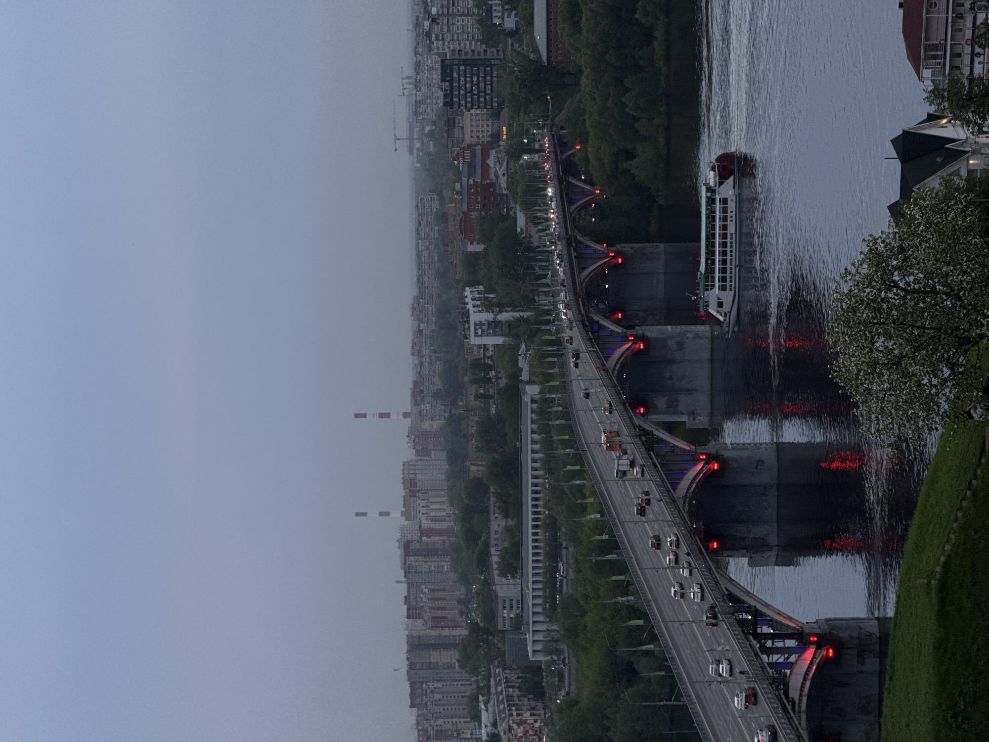 An elevated aerial perspective shows a bridge spanning a wide river. The bridge is lit with red lights and has cars driving across it. A large boat is moving along the river. The river reflects the lights and the boat. Tall buildings are visible on both sides of the river, with smokestacks rising in the distance on the left. Trees line the riverbanks and separate the city from the river. The sky is overcast. This view is the Kanavinsky Bridge in Nizhny Novgorod, Russia.