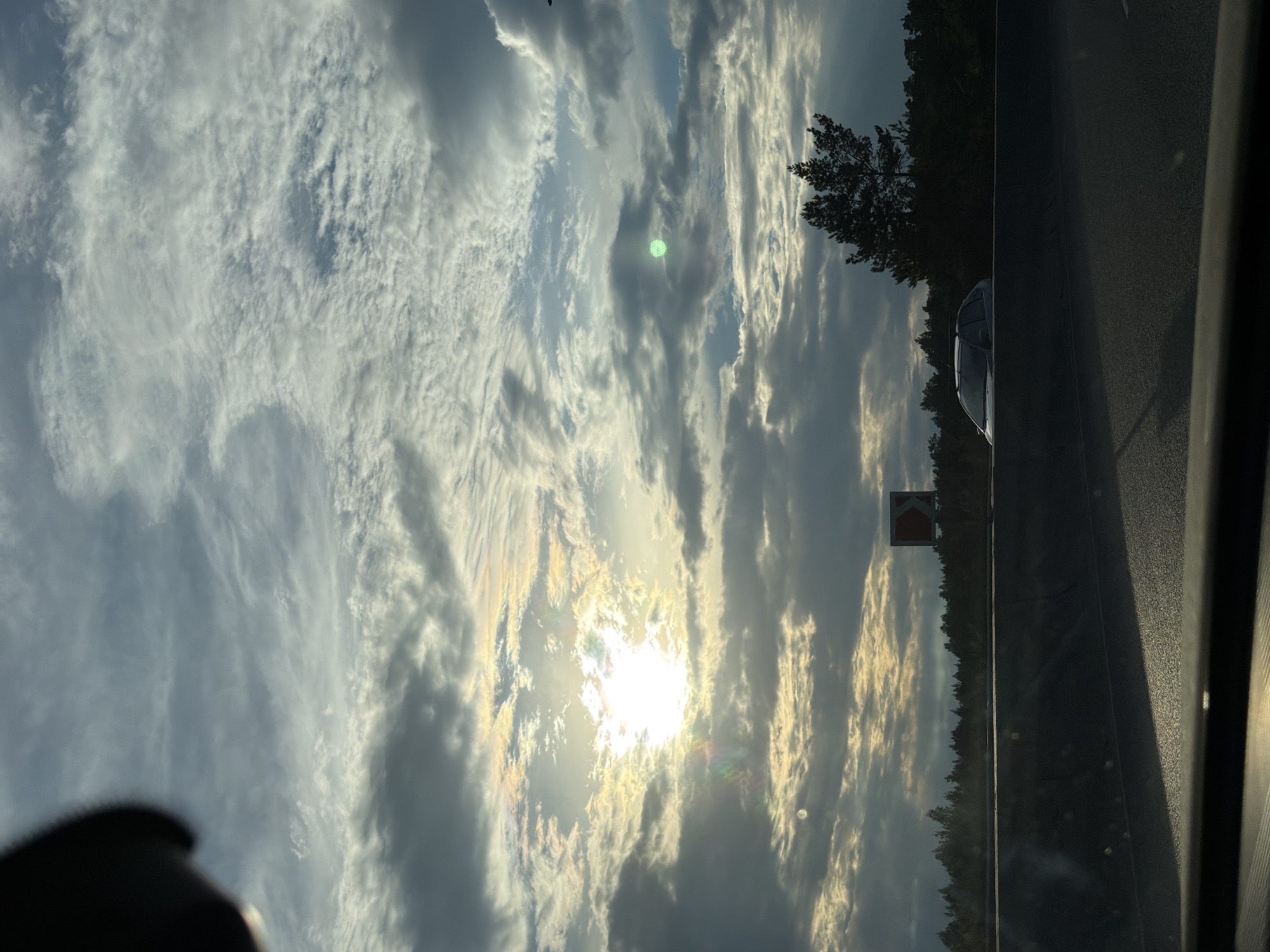 The image depicts a scenic view from a moving vehicle, likely on a highway, as indicated by the presence of a road and a car. The perspective suggests the photo was taken looking upwards and outwards. The sky is filled with dramatic clouds, illuminated by the sun which is partially obscured by them. The sun's rays create a dazzling effect, with visible lens flares and a rainbow-like halo. Some clouds are wispy and white, while others are darker and more imposing, creating a dynamic contrast.
To the right of the frame, a dark line of trees forms a treeline, suggesting a rural or natural setting adjacent to the road. A single tree with a distinctive shape stands out against the sky. Further along the road, a traffic sign with an orange chevron is visible. A silver car is also seen on the road, slightly ahead of the viewer's vehicle. The overall impression is one of a journey during a striking sunset or sunrise, with the clouds and light contributing to a dramatic and perhaps awe-inspiring atmosphere. The time of day is likely early morning or late afternoon given the low angle of the sun and the quality of light. The weather appears to be partly cloudy, with breaks of sunlight. The location context of Perm, Russia, implies this is a view experienced during travel within that region.