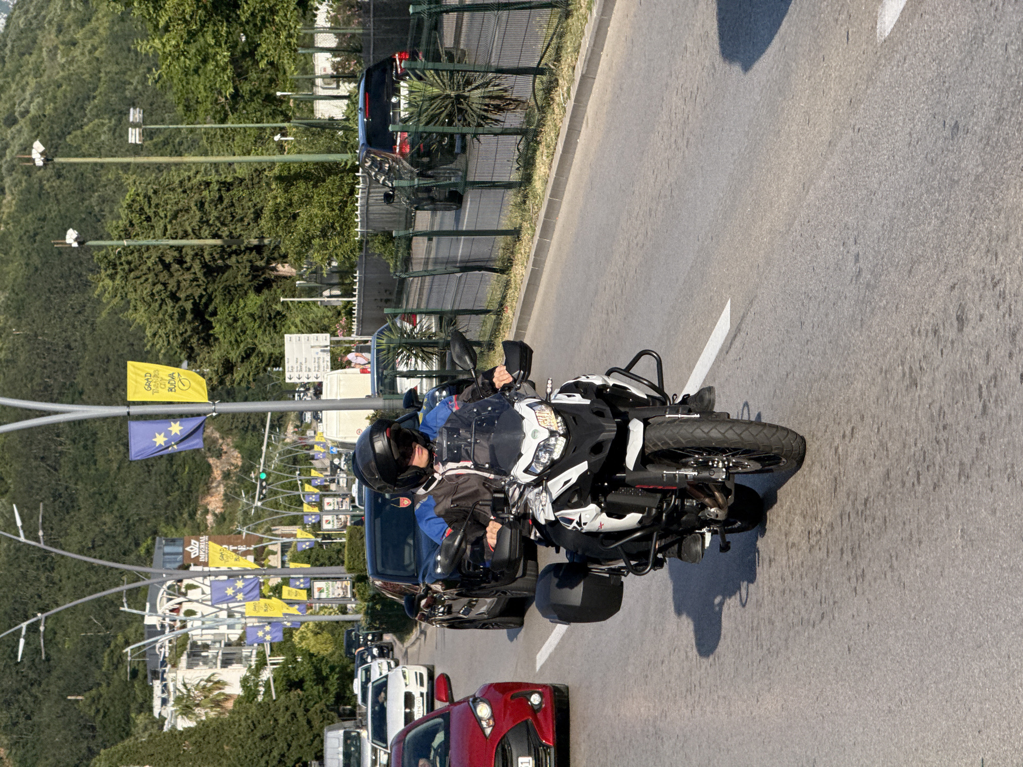 A motorcyclist wearing a black helmet and a blue and black jacket rides a white and black adventure motorcycle down a paved road in Budva, Montenegro. The motorcyclist is in the foreground, and the motorcycle has saddlebags on either side. In the background, other vehicles, including a red car and a black SUV, are visible on the road. Tall trees and buildings line the side of the road, and a hillside covered in vegetation is in the far background. Several flags are hanging from lampposts.