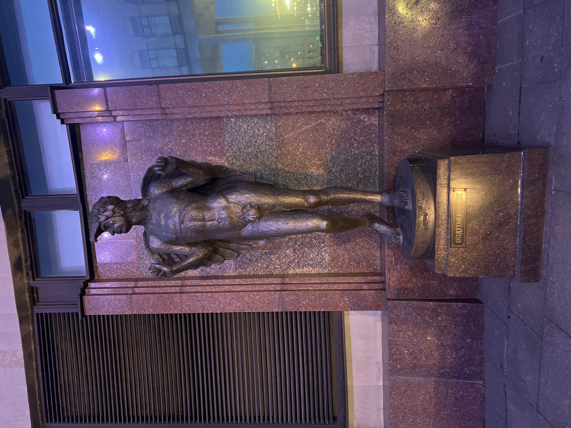 The image shows a bronze statue of a young man standing on a pedestal in front of a building. The statue is of a naked male figure wearing a helmet and has wings attached to its back. The man is holding a bundle of something over his shoulders. The building behind the statue has a large louvered window on the left and a modern window on the right, reflecting the surrounding city lights. The pedestal of the statue has an inscription on it. The building's facade is made of granite with red and grey speckles. The statue is illuminated by artificial lights, giving it a warm glow. The ground in front of the statue is paved with square stones. The overall scene suggests an outdoor public space, possibly in a city, at night. The inscription on the pedestal is in Cyrillic and appears to read "ВОЗДУШНАЯ", possibly part of a longer title related to air or flight. The statue is likely located in Saint Petersburg, Russia.