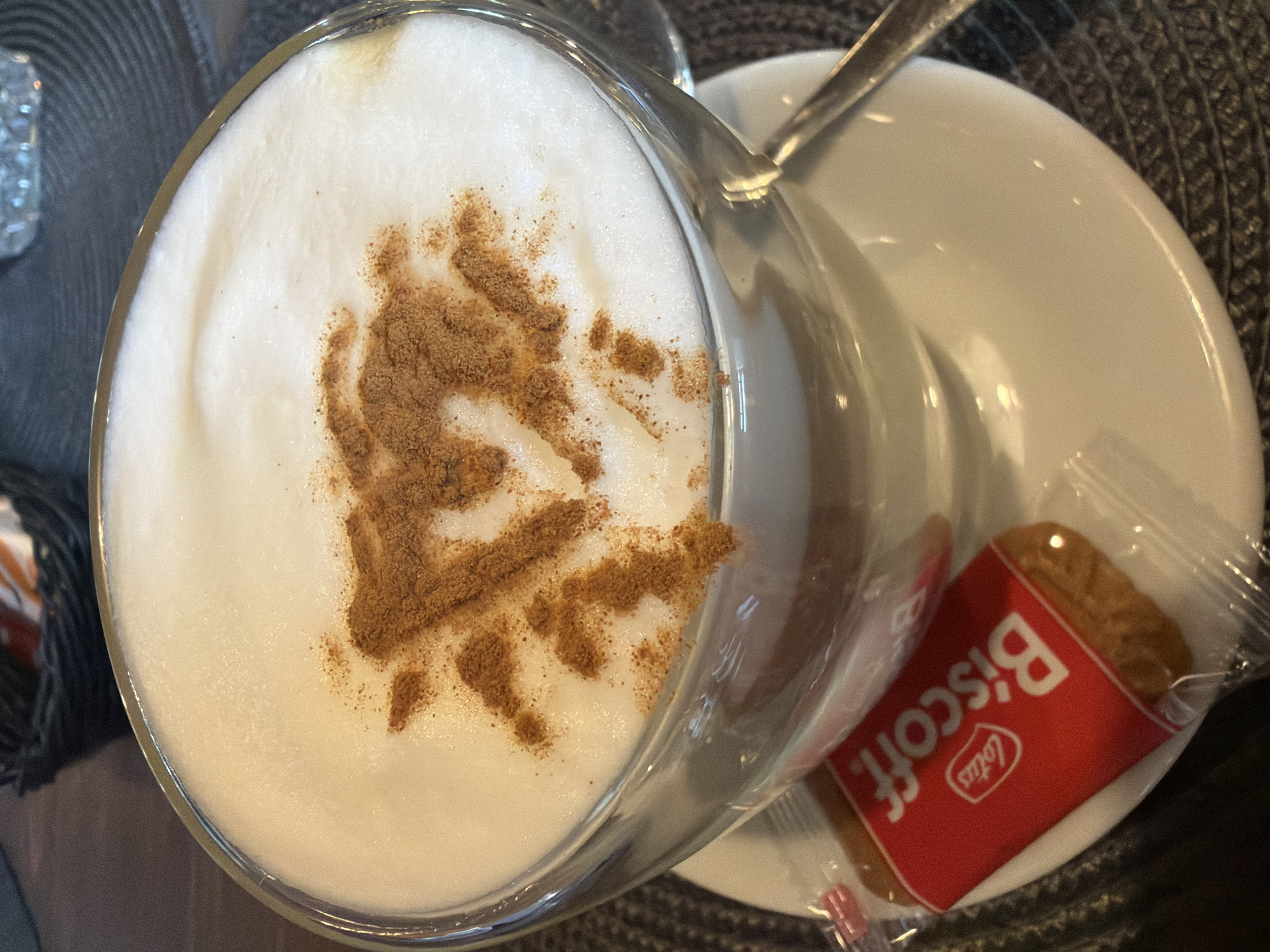 The image shows a close-up, overhead view of a cappuccino with cinnamon dusted on top, resembling a fir tree. The coffee is served in a clear glass mug, which is placed on a white saucer. A spoon rests on the saucer, partially submerged in the coffee. Beside the mug, a single Biscoff cookie in its red wrapper is visible, also on the saucer. The setting appears to be a cafe or restaurant, indicated by the woven placemat beneath the saucer and the subtle background details. The lighting suggests it could be daytime. The overall mood is relaxed and inviting, typical of enjoying a warm beverage. Visible text on the cookie wrapper reads "Lotus Biscoff". The location is Madona, Latvia.