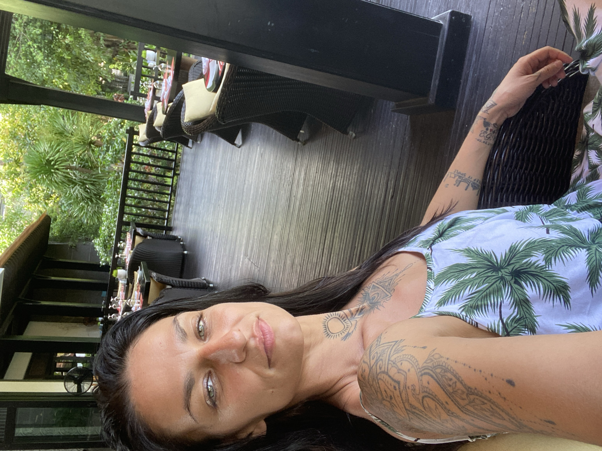 A woman with long, dark hair and green eyes is pictured from the chest up, facing forward and slightly to her right. She has tattoos on her neck, shoulder, and arm. She is wearing a white tank top with a palm tree pattern. The background shows an outdoor seating area of a restaurant with wooden tables and chairs, set up on a wooden deck. Behind the seating area, there is a black railing and lush green foliage, suggesting a tropical environment. The lighting indicates it is daytime, possibly late afternoon, with dappled sunlight filtering through the trees. The location is described as Phu Thuy Ward, Vietnam.