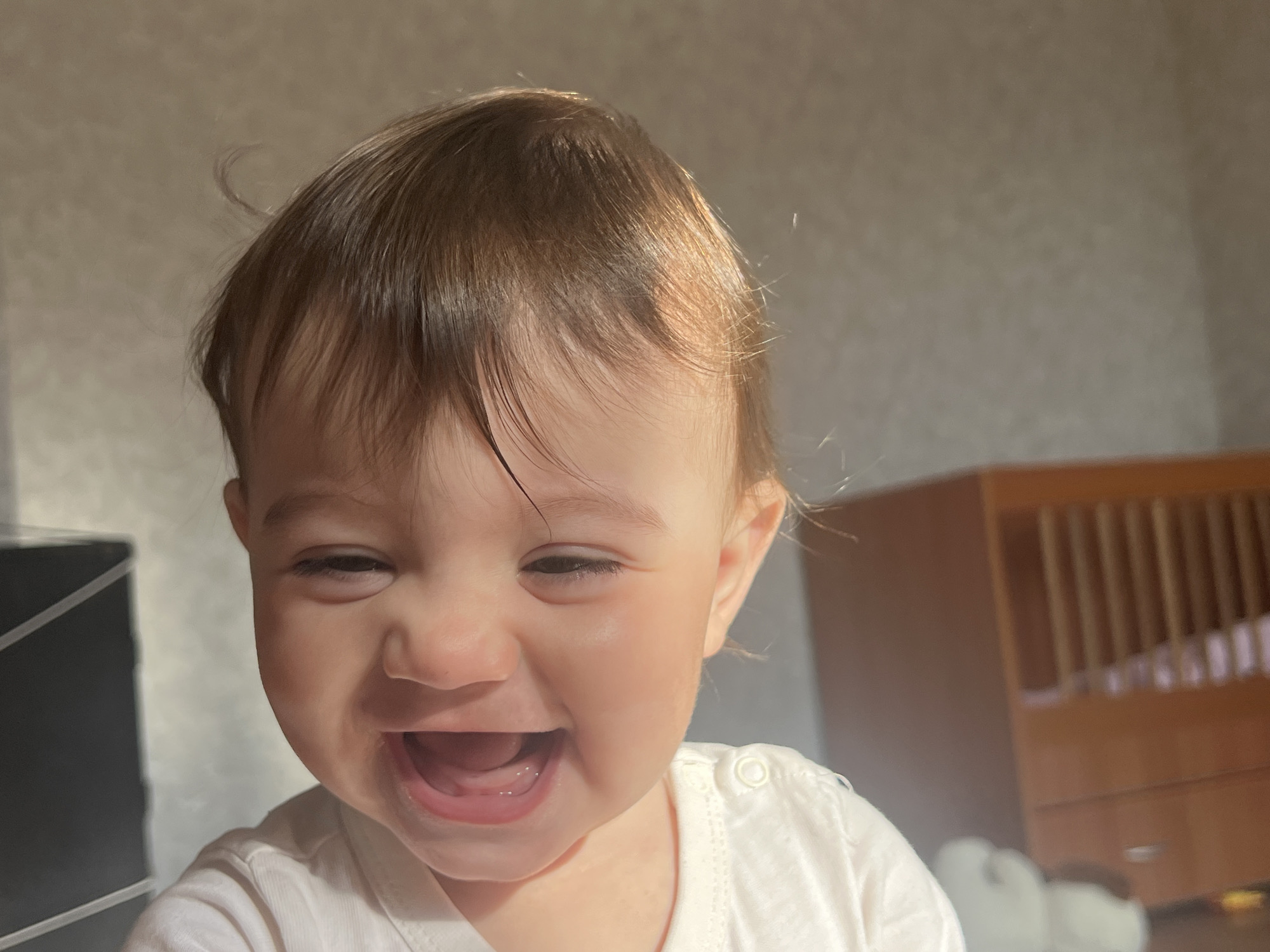 The image displays a close-up, head-and-shoulders portrait of an infant, approximately 6-18 months old, captured indoors within a residential setting in the city of Elena, Bulgaria. The infant, possessing light brown hair and fair skin, is depicted in a state of laughter, with eyes squinted shut, cheeks raised, and mouth wide open, revealing a pink tongue. The subject is wearing a plain white garment, likely a top or onesie, with a visible white snap button on the right shoulder. Strong, warm directional lighting, consistent with natural sunlight, illuminates the right side of the infant's face and hair, creating highlights. The background on the left features a dark, rectangular object with lighter linear elements. To the right, a light-colored wooden crib with vertical slats is partially visible, along with indistinct, light-colored objects on the floor. The wall behind the infant is a light neutral tone with a subtle, uniform texture.