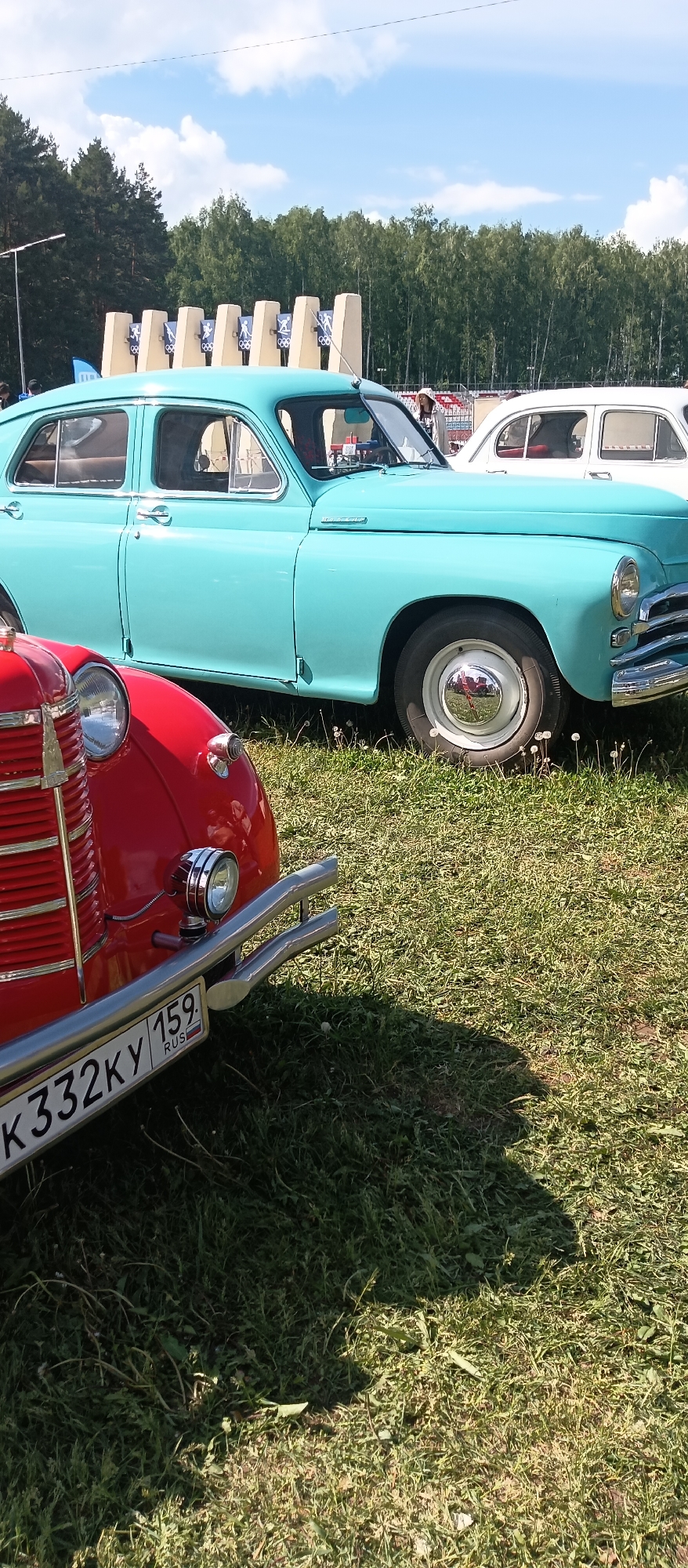 The image shows a sunny outdoor scene featuring two vintage cars parked on a grassy area. The foreground displays the front of a red car with a visible license plate that reads "K332KY 159 RUS." Beside the red car is a blue car with a chrome hubcap. In the background, an Olympic monument stands in front of a line of trees, and a white car is visible. The location appears to be a park or field in an urban or suburban area.