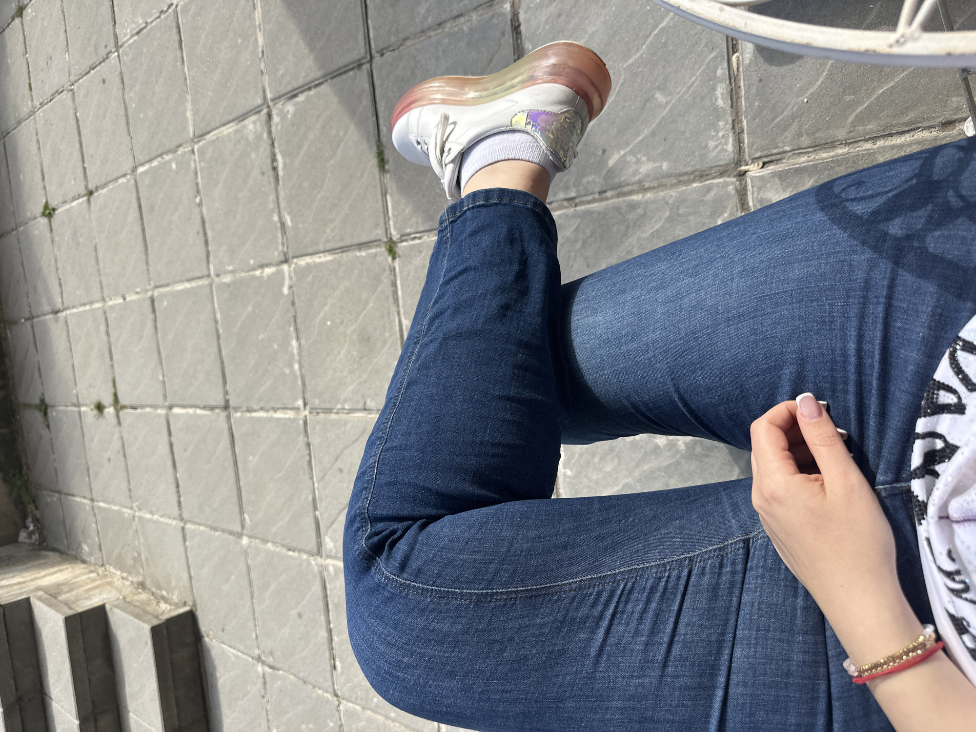 The image captures a top-down view of a person seated outdoors, primarily showing their legs, one arm, and a hand. The person is wearing dark blue denim jeans, white ankle socks, and white athletic shoes with distinctive iridescent accents and a translucent, reddish-pink sole. One leg is bent, with the foot slightly raised, showcasing the shoe. The other leg is also bent, with the arm resting on the thigh. The person's left hand, adorned with French manicured nails and a few beaded and red string bracelets on the wrist, is gently placed on their leg.
The scene is set on a paved surface made of square grey tiles, which appear somewhat worn. Patches of small green weeds are visible in some of the tile gaps. Bright sunlight casts sharp, distinct shadows across the ground, notably from the person's body and from a white, possibly metal, chair or table leg in the upper right corner of the frame. In the lower-left corner, a series of grey concrete steps descend, suggesting a change in ground level or an entrance/exit point. The overall impression is a sunny day in an urban or semi-urban outdoor setting, possibly a cafe or public seating area in Sofia, Bulgaria. No visible text is present in the image.