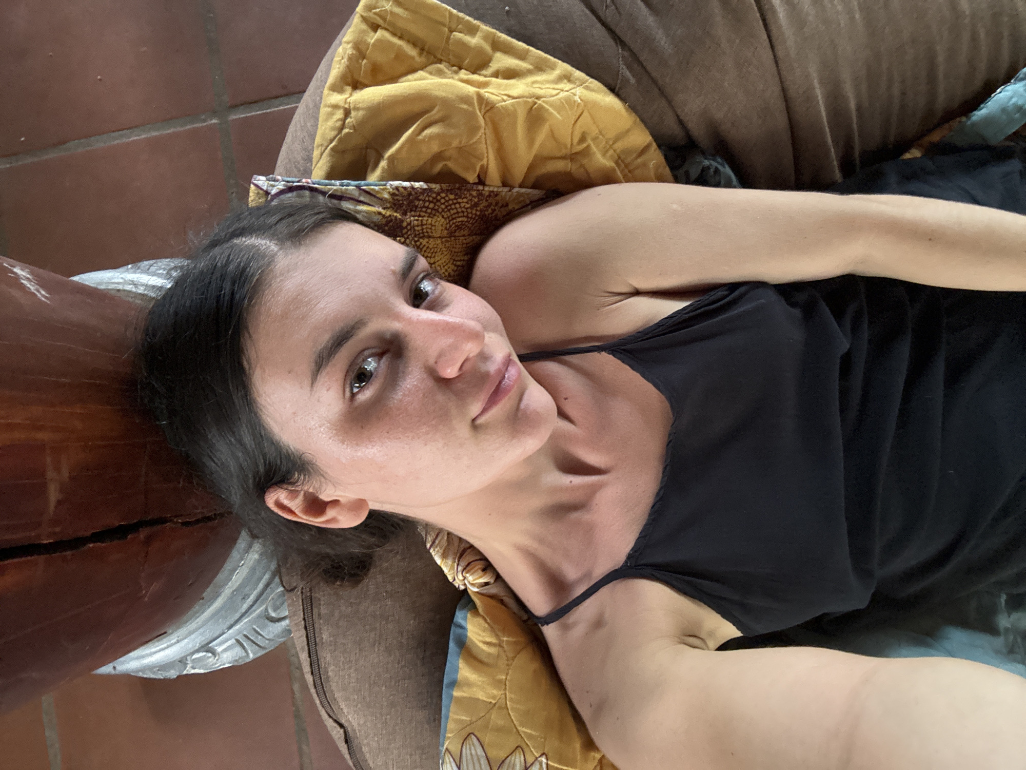 A woman lies on a beanbag chair, her head propped on a yellow pillow with a floral pattern. She is wearing a black tank top and looking directly at the camera with a slight smile. The background shows tiled flooring and the base of a large, wooden object, possibly a mortar or a decorative pillar. The scene appears to be indoors, or at least under a covered area, given the tiled floor and the presence of soft furnishings. The lighting suggests it is daytime. The woman's hair is dark and pulled back. Her skin is slightly flushed, and there are visible pores and some light blemishes on her face. Her eyes are dark brown and open. She is lying on her back, with her right arm bent and resting near her head, and her left arm extended downwards, out of frame. The overall atmosphere is casual and relaxed. The image was taken in Mũi Né, Vietnam, a coastal resort town known for its sand dunes and fishing activities.