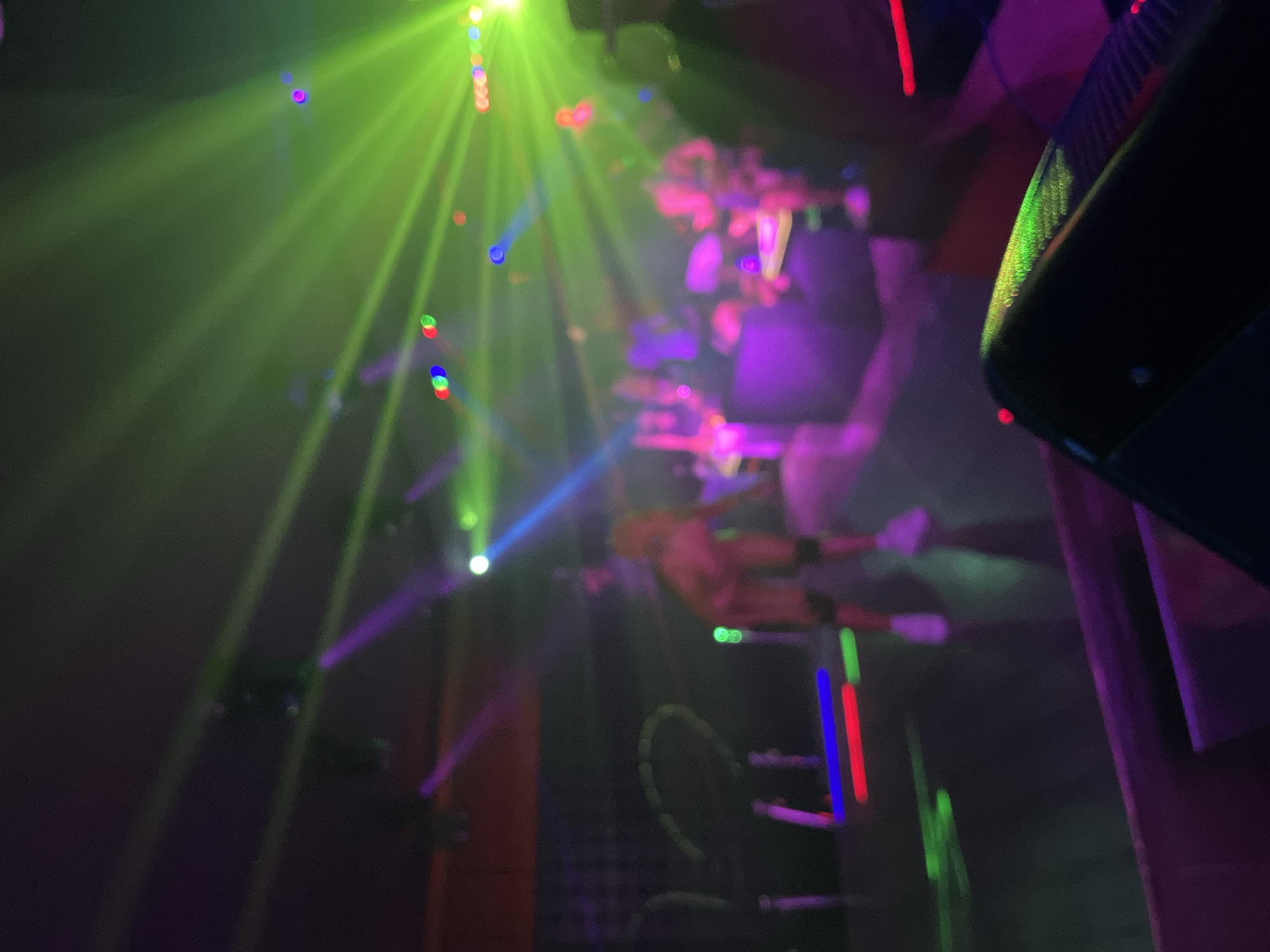 This is a photograph taken at night, likely indoors, with vibrant colored lights illuminating the scene. Green and purple lights dominate the frame, creating beams and casting a hazy, atmospheric glow. In the center of the image, a person appears to be suspended or performing an acrobatic move, dressed in what looks like a light-colored, minimal outfit. They are blurred due to motion and the low light conditions.
Surrounding the performer, there are various objects and people, all appearing indistinct. Some are holding glowing sticks in red, blue, and green. A large black object, possibly a speaker or equipment, is visible in the foreground on the right. The overall setting suggests a club, concert, or a performance venue. The mood is energetic and festive, conveyed through the dynamic lighting and the implied activity. There is no visible text in the image. The location context of Howayi, Nigeria, does not provide specific visual cues within the photograph itself, but indicates the geographical setting of this event.