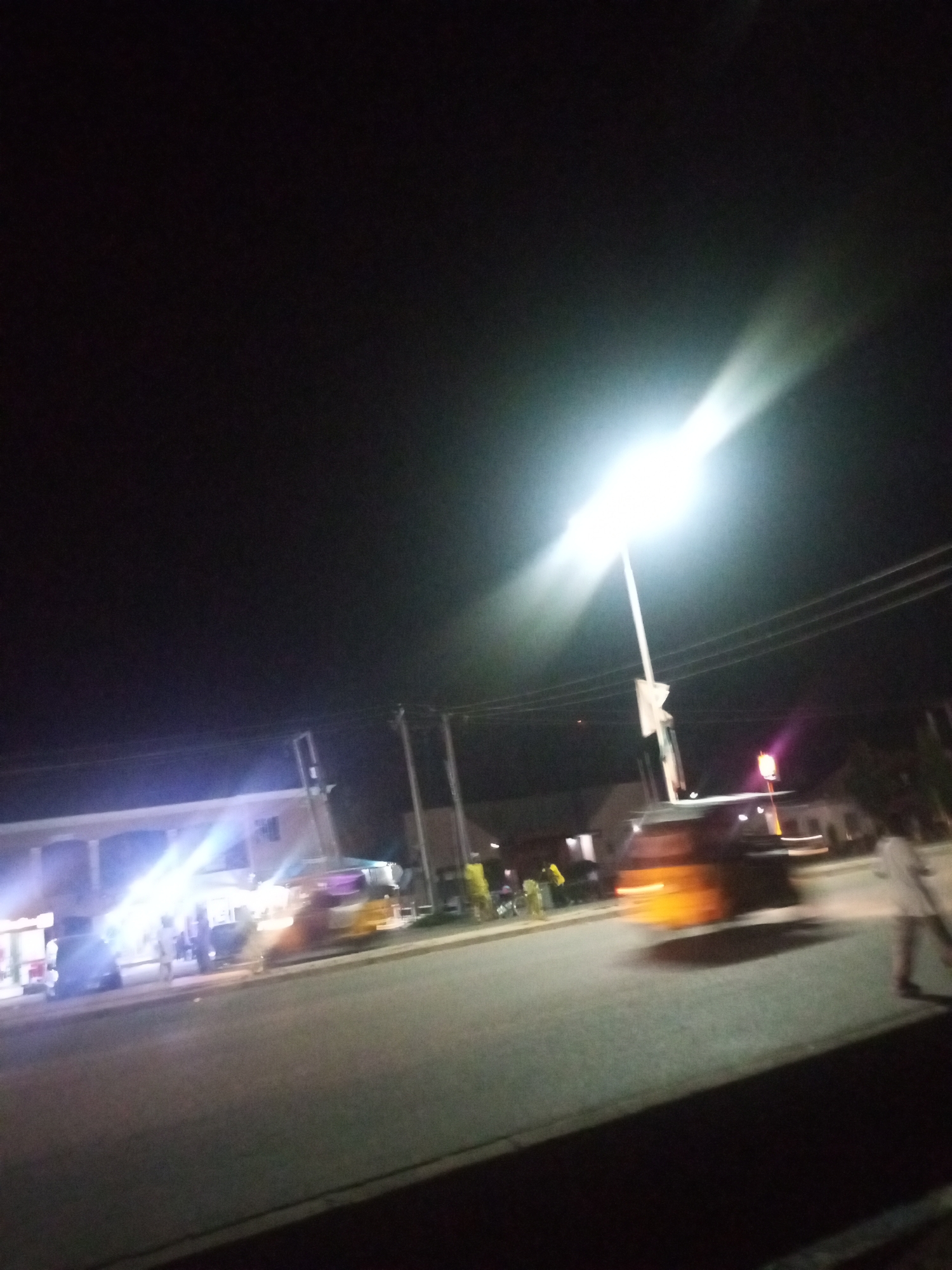 The image depicts a street scene at night, possibly in the city of Unknown. A bright street light dominates the upper right portion of the frame, casting a strong beam across the scene. The road is visible in the foreground, with a vehicle, possibly a tuk-tuk or auto-rickshaw, in motion on the right side, its headlights and body blurred due to movement. On the left side of the road, there appears to be a building with bright lights illuminating its facade. Several indistinct figures of people are present, some near the building and one on the far right walking away from the camera. Utility poles and wires are visible against the dark night sky. The overall impression is that of a busy street corner or marketplace at night, with artificial lighting creating a dramatic contrast with the darkness.