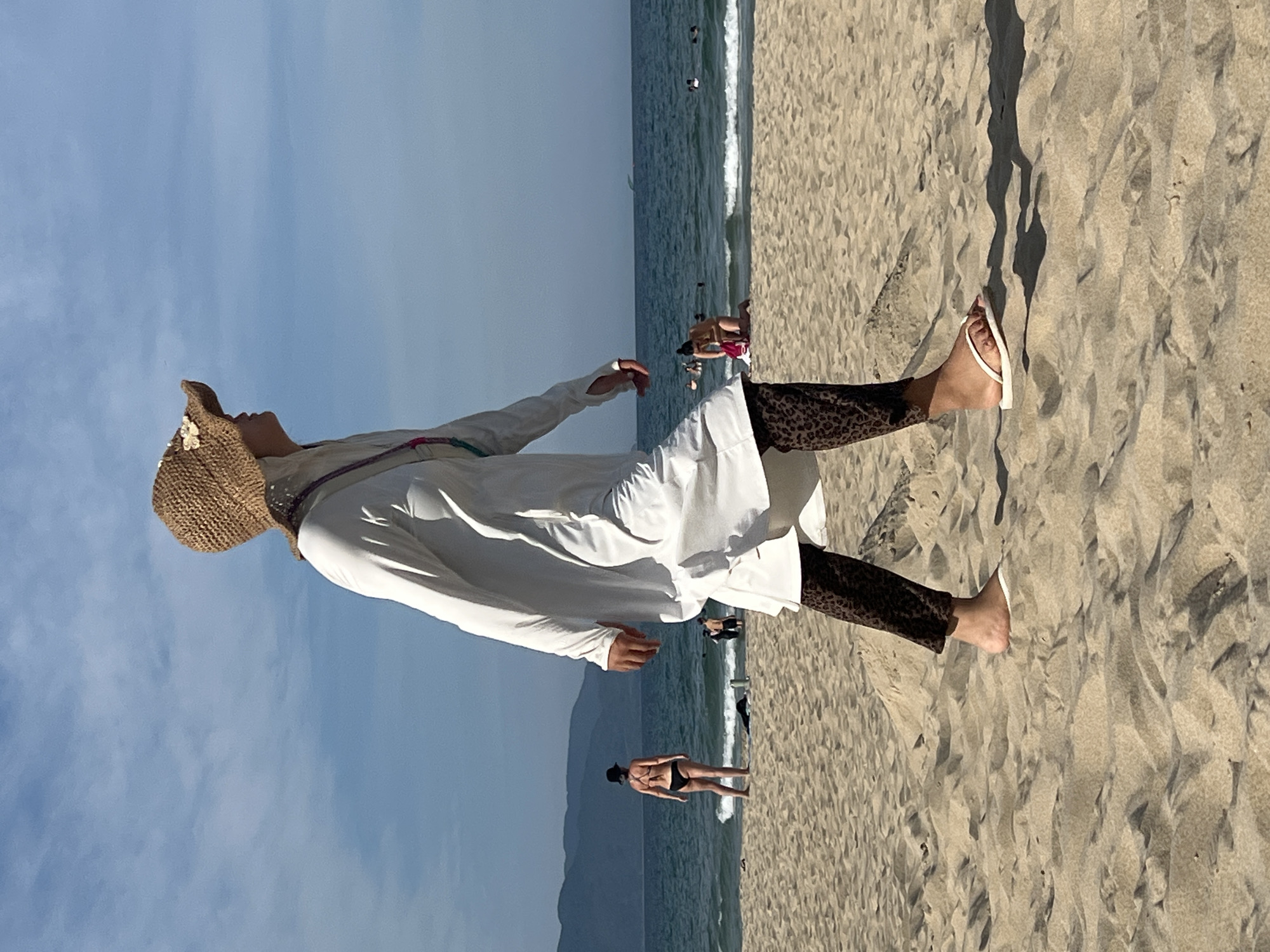 A woman wearing a white long-sleeved dress, a straw hat adorned with flowers, and leopard-print leggings walks on a sandy beach. She is wearing white flip-flops. In the background, the ocean meets the horizon under a blue sky with scattered clouds. Several other people are visible on the beach and in the water. Some are swimming, while others are relaxing on the sand. In the distance, mountains form a backdrop to the scene. This appears to be a typical scene at a beach in Đà Nẵng, Vietnam, a popular coastal city known for its sandy shores and clear waters.