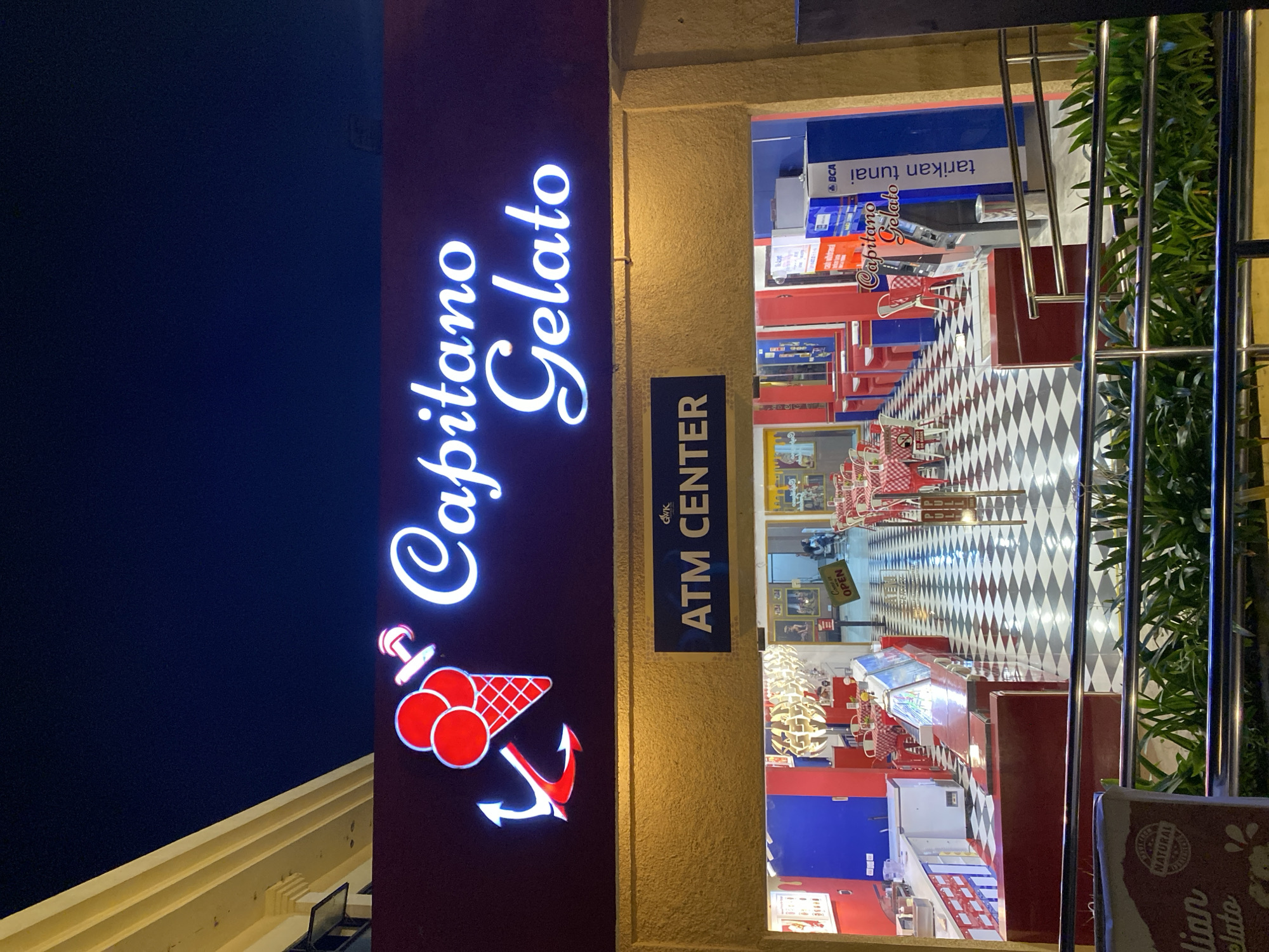The image shows the exterior of a Capitano Gelato shop and an ATM center at night in Kuta Selatan, Indonesia. The shop's sign, illuminated with neon lights, features a red ice cream cone with an anchor. Below the sign, a blue banner reads "ATM CENTER." The entrance to the shop reveals a checkered floor, red tables with white chairs, and an ice cream display case. An ATM machine with "BCA" branding is visible to the right of the entrance. The surrounding environment is dark, indicating it is nighttime.