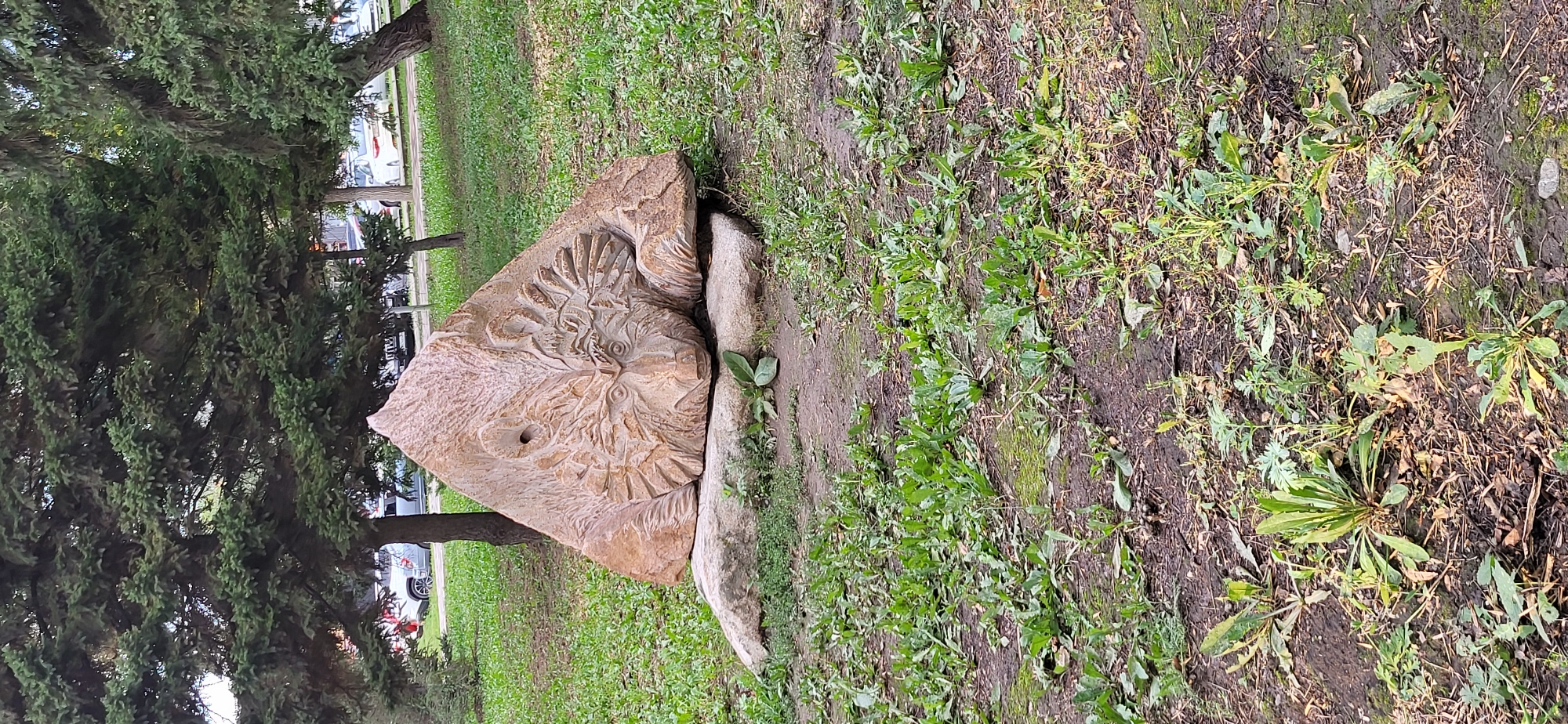 The image shows a large, carved stone sculpture resembling an owl's face, placed on a grassy area in what appears to be a park or public space. The sculpture is triangular in shape and has intricate carvings of feathers and eyes. A small, round hole is visible on the left side of the sculpture, resembling an owl's pupil. The sculpture is supported by two flat, light-colored stones. In the background, trees with green foliage are visible, along with a road and parked cars. The ground around the sculpture is covered in grass and dirt. The image is taken from a slightly low angle, focusing on the sculpture. This sculpture is located in the park of the city of Oskemen, Kazakhstan.