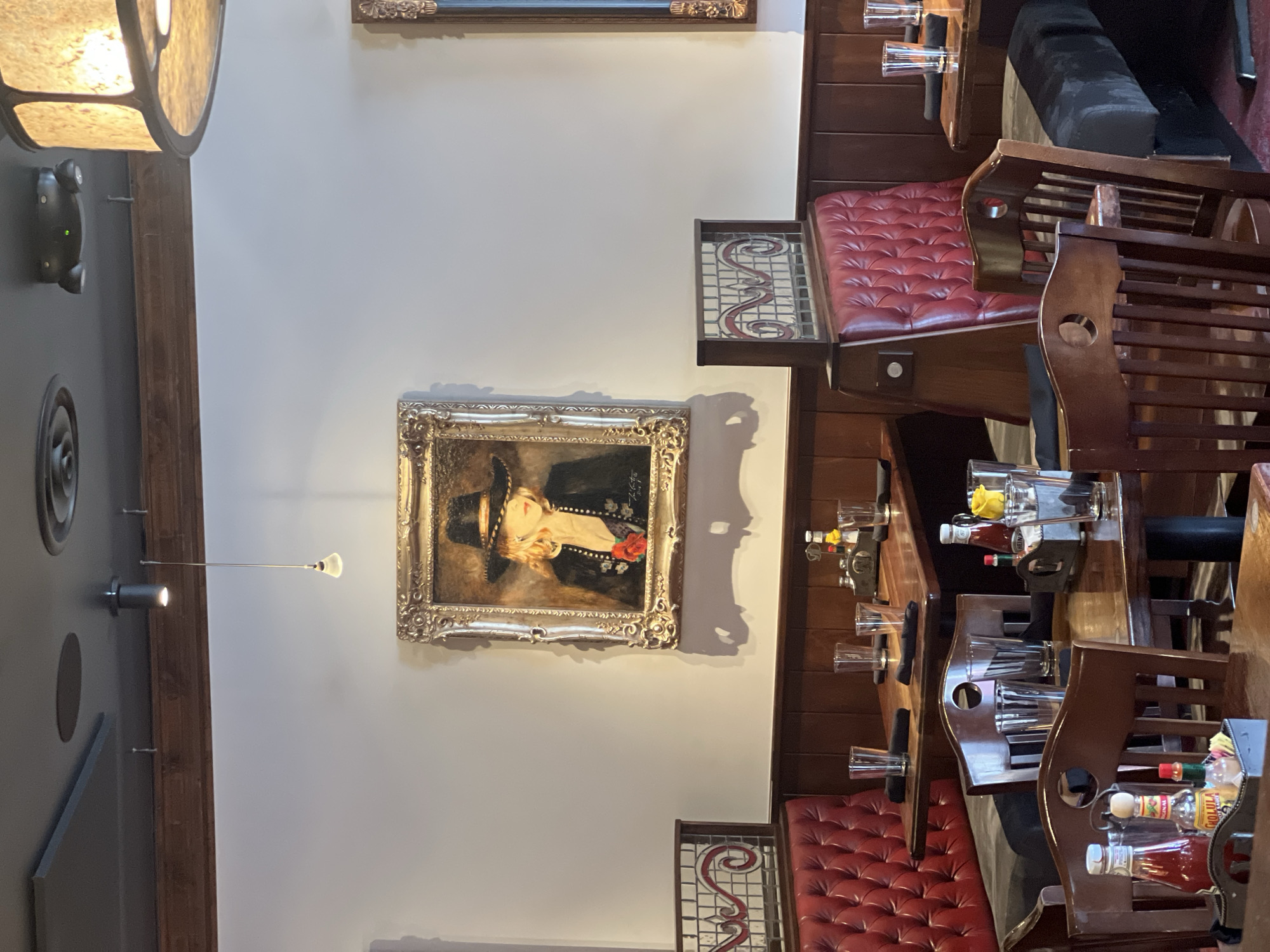The image shows the interior of a restaurant in Jackson, United States of America. The restaurant has a warm and inviting atmosphere with dark wood paneling, red leather booths, and wooden chairs. A painting of a woman in a black hat and jacket with a red rose is displayed on the wall, adding a touch of elegance to the space. The tables are set with glasses, napkins, and condiments, indicating that the restaurant is open for business. The lighting is dim and ambient, creating a cozy and intimate dining experience.