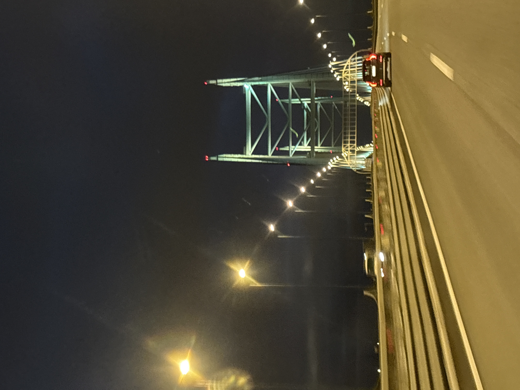 The image captures a nighttime view of a bridge, likely a highway overpass, with a car driving away from the camera. The bridge structure is illuminated, and streetlights line the roadway. The sky is dark, suggesting late evening or night. The location is identified as Unknown, Russia. The scene appears to be on a clear night, with no rain or fog evident. The car in the foreground is a dark-colored SUV, its taillights are illuminated. The roadway has multiple lanes, and the bridge itself is a prominent architectural feature in the background.