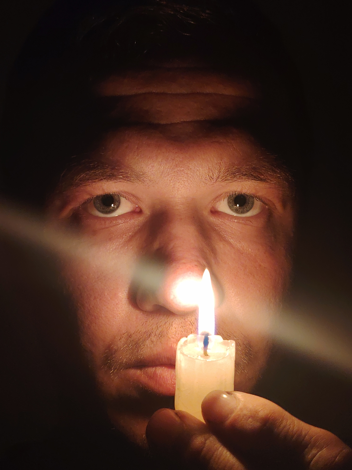 The image is a close-up, low-light, portrait of a person's face, illuminated by a lit candle held in their hand. The person's eyes, nose, and mouth are visible, with the flame of the candle positioned directly in front of their nose and mouth. The background is dark, with the light from the candle casting a glow on the person's skin and creating a bright flare across their face. The person's hand, holding the candle, is also visible in the foreground. There is no indication of the city of Chaikovsky, Russia, or any other specific location within the image.