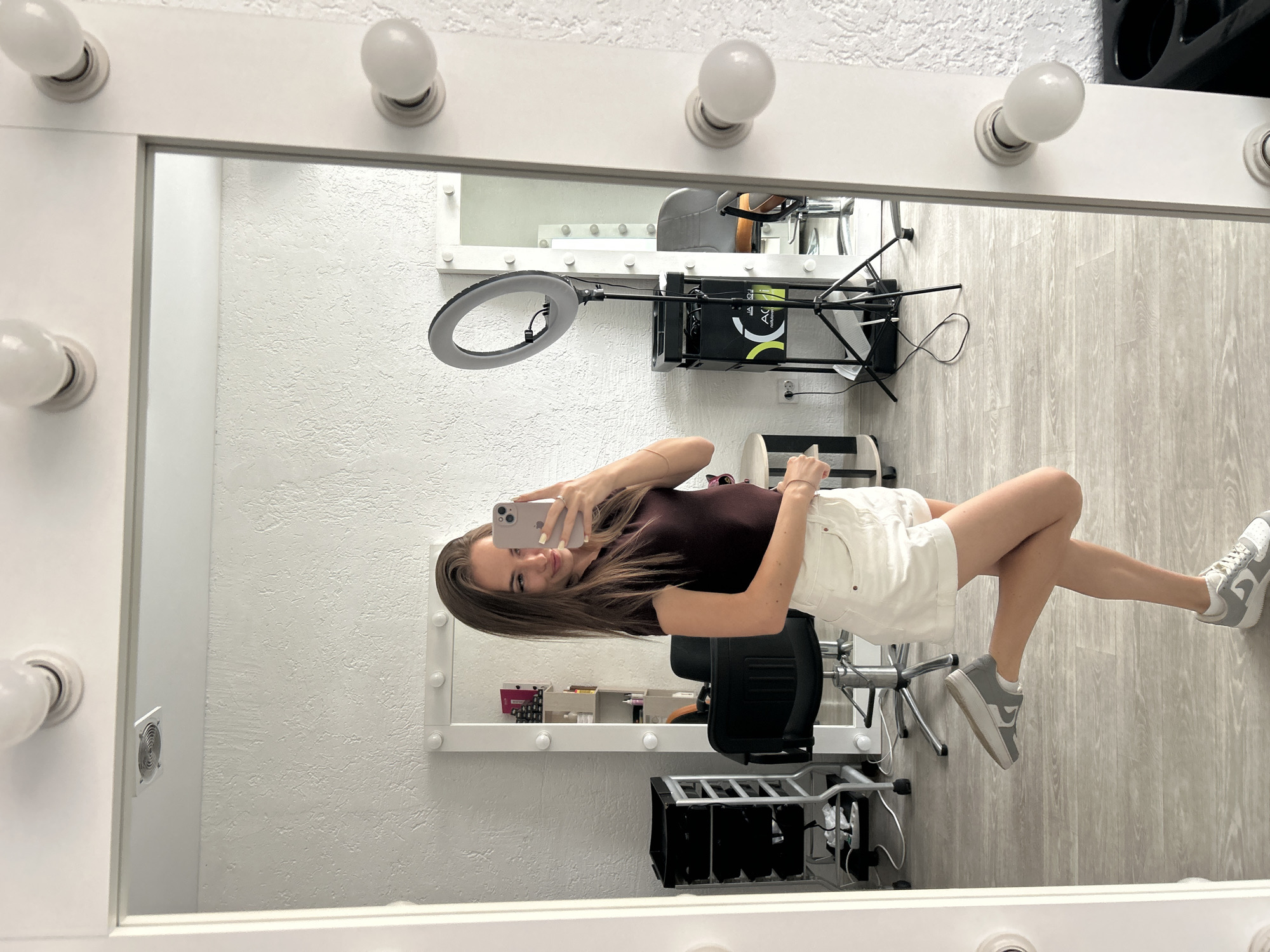 A woman with long brown hair is standing in front of a large, brightly lit mirror, taking a selfie. She is wearing a dark maroon t-shirt and white denim shorts, with gray and white sneakers. Her left leg is bent at the knee and slightly lifted behind her. The mirror has a white frame with numerous light bulbs around its edge. Reflected in the mirror is a room that appears to be a salon or a dressing area. Visible in the reflection are salon chairs, a rolling cart with shelves, a ring light on a tripod, and shelving units with various products. The walls are textured and painted white, and the floor is light gray wood. The overall lighting is bright, coming from the mirror's bulbs and overhead sources.